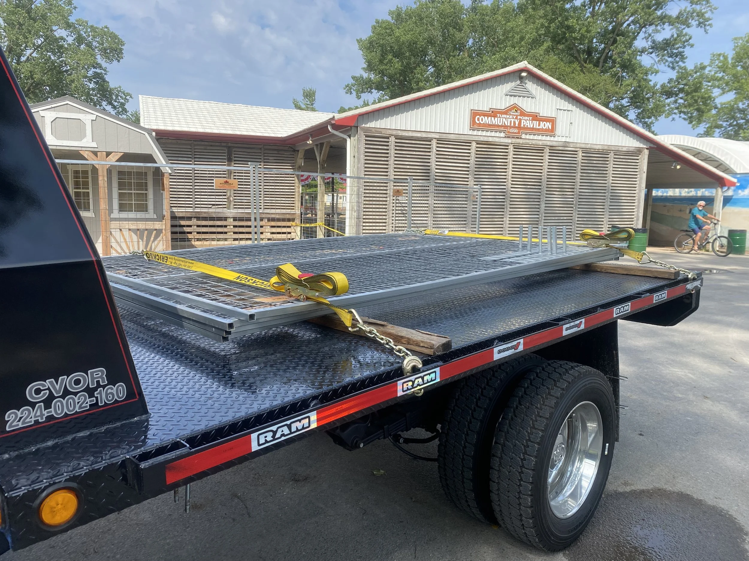 Flatbed truck with metal gate on it, parked in front of community pavilion with a person riding a bicycle in the background, sign reading 'Turkey Point Community Pavilion' on the pavilion building