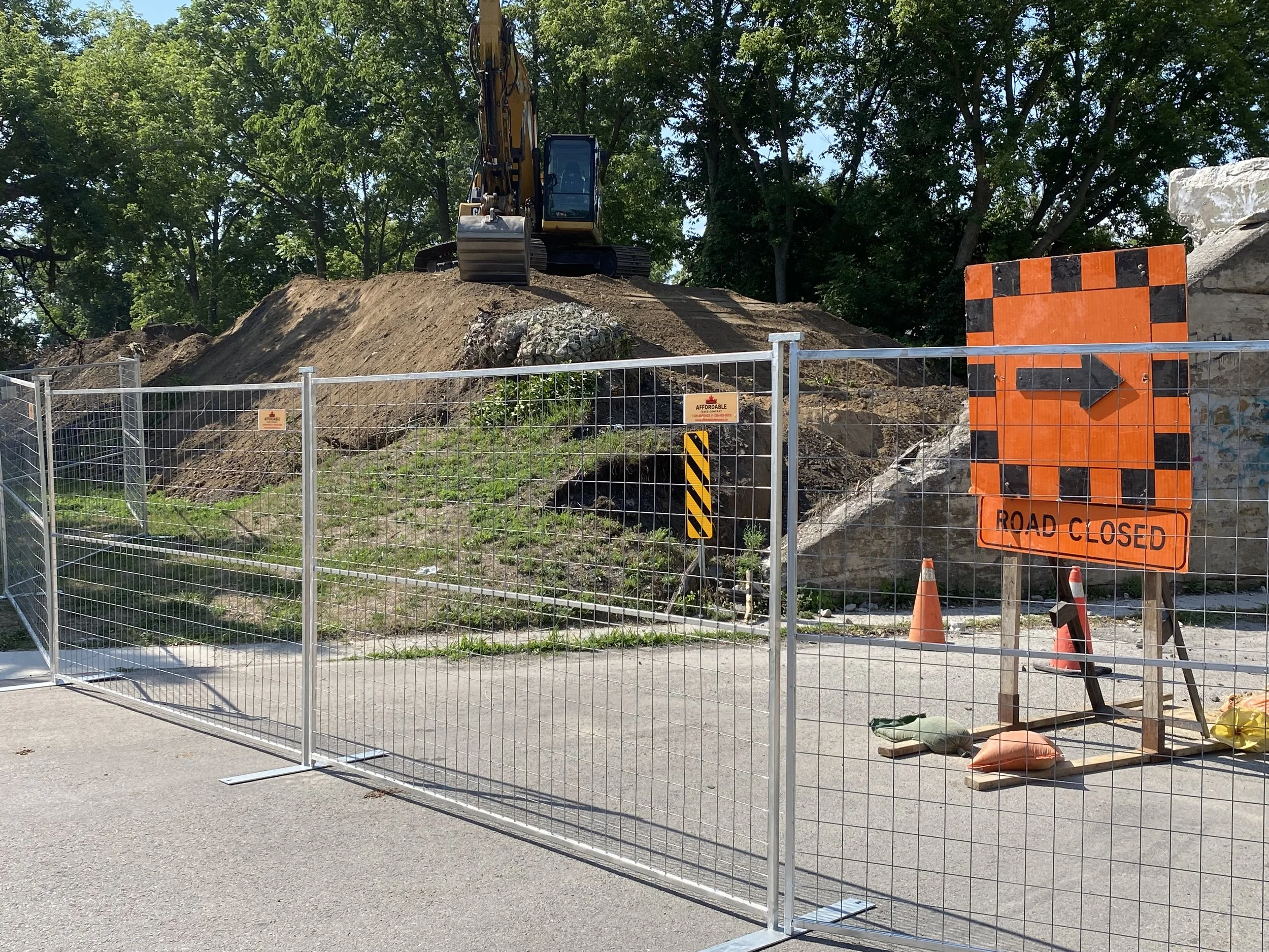 Construction site with a sign that reads 'Road Closed' and a fence blocking the street. There is a large mound of dirt with an excavator on top, surrounded by trees and clear blue sky.