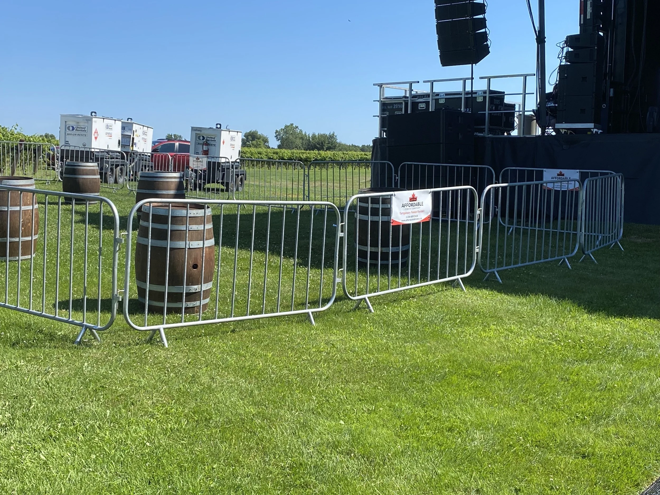 Outdoor stage area with barricades, wooden barrels, and equipment, set on a grassy field on a sunny day.