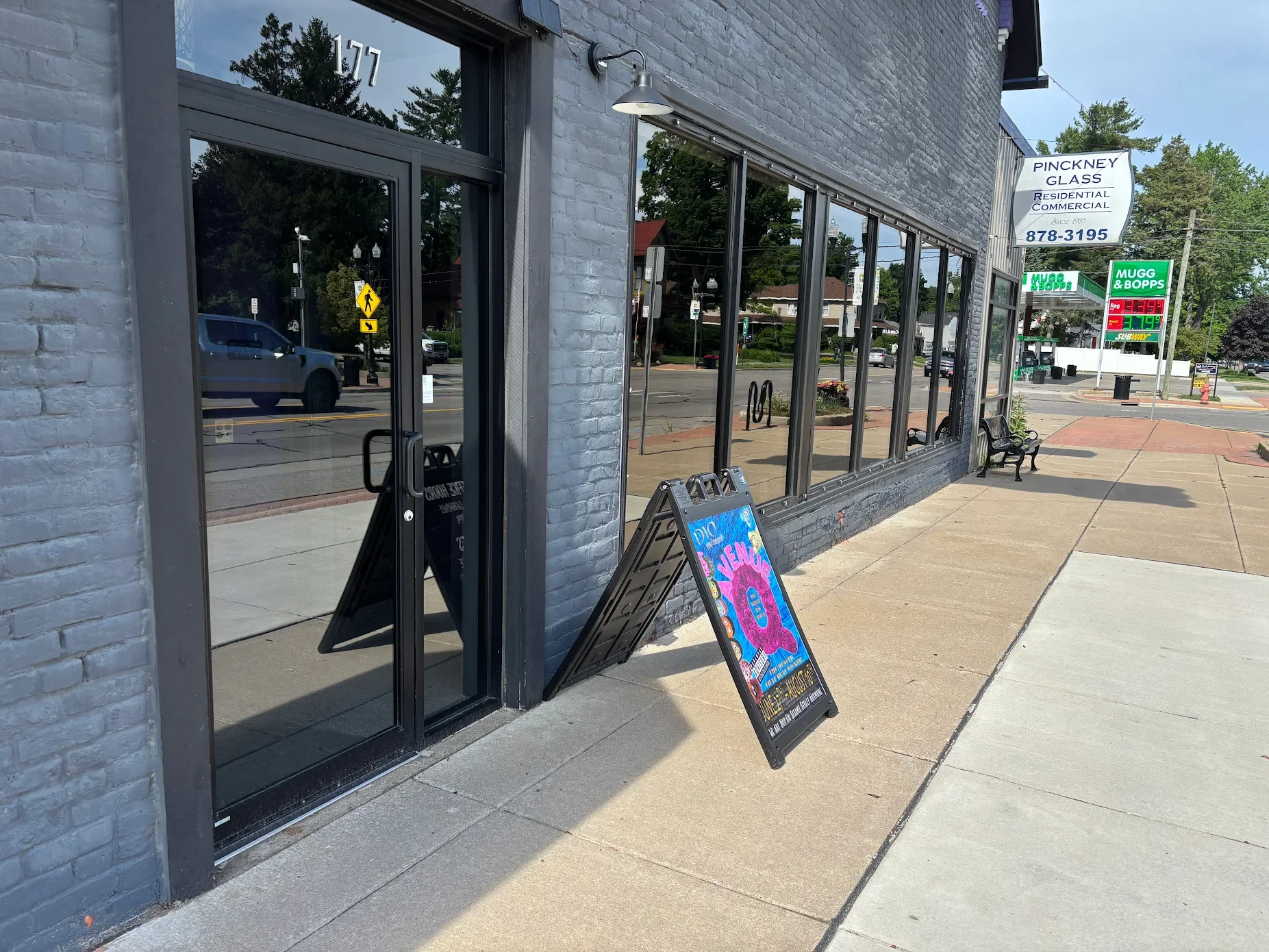Empty sidewalk outside a gray brick building with glass windows and city street reflection, featuring a sidewalk signboard advertising an event, benches, and a parking lot in the background.