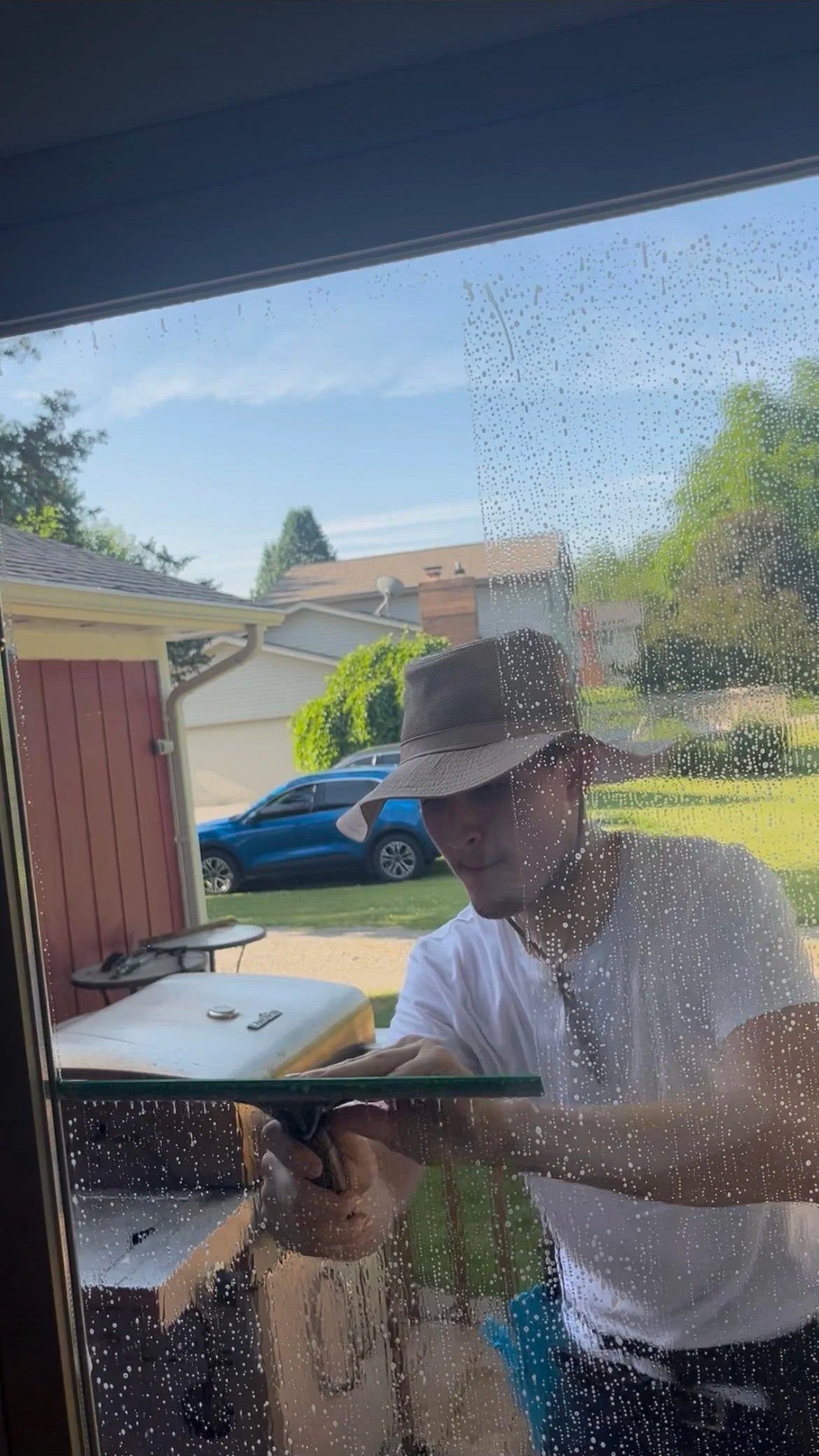 Panther Pro Cleaning wearing a wide-brimmed hat cleaning a glass door with water droplets, outdoors with a blue car and houses in the background.