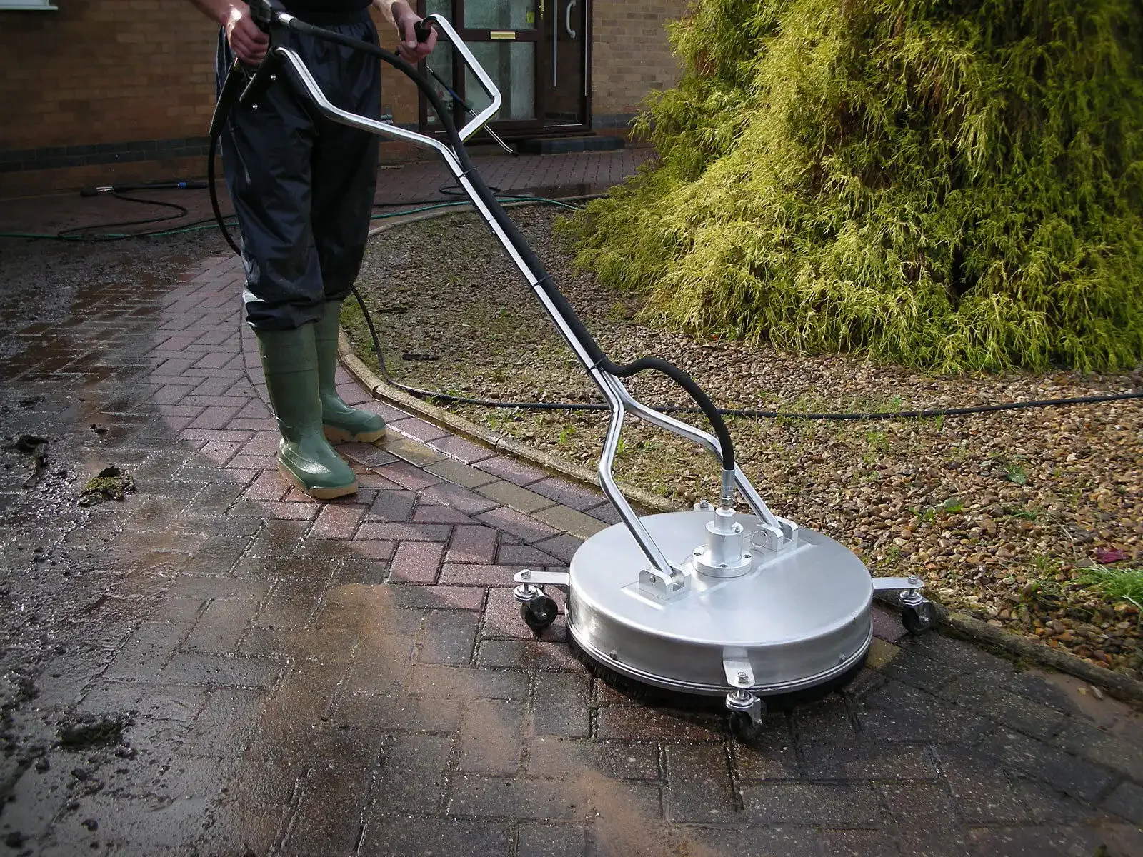 Person in green boots operating a large, professional-grade sidewalk cleaning machine on a brick driveway during rainy weather.