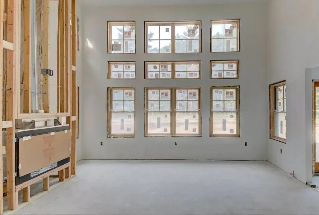 Interior of a house under construction with multiple window openings on a wall, exposed wooden framing on the left, and white walls.