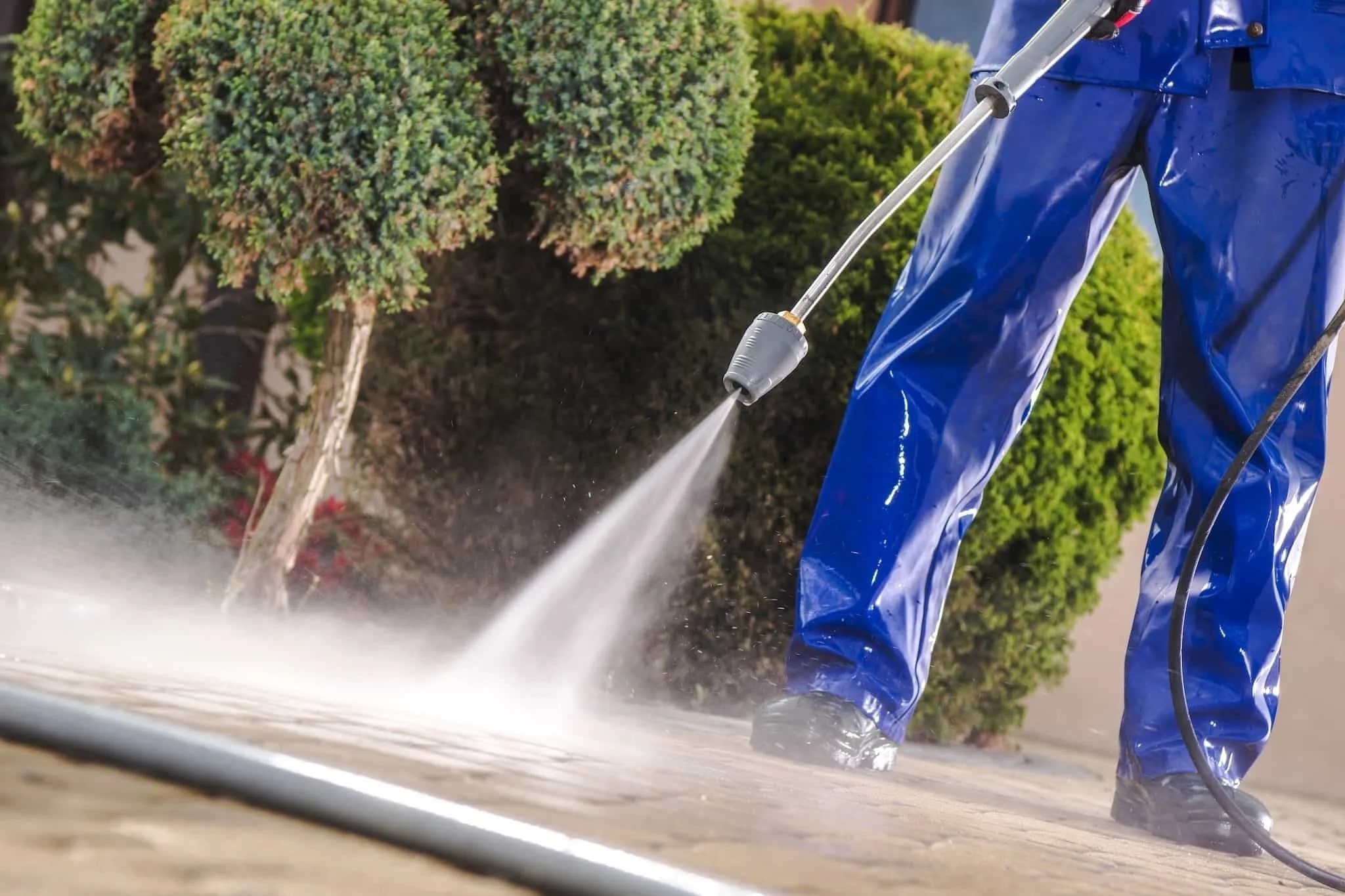 Person in blue waterproof pants and shoes pressure washing a sidewalk.