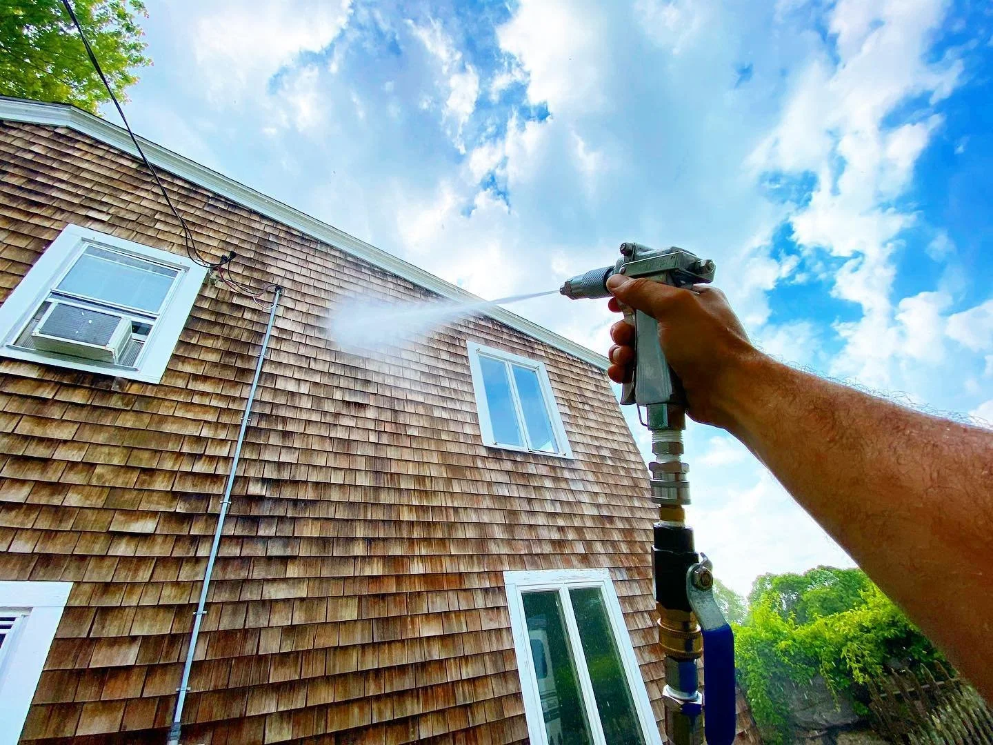 A person using a garden pressure washer to clean the exterior wall of a house with brown wooden shingles, windows, a downspout, and a clear blue sky with clouds in the background.