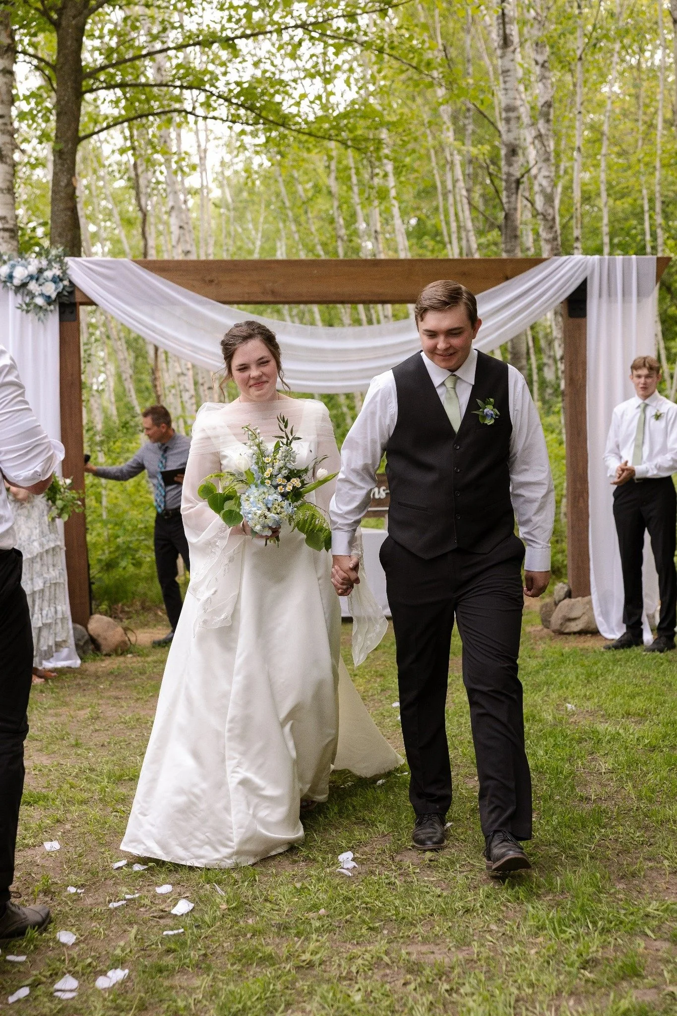 A bride and groom walk hand in hand outdoors during a wedding ceremony, surrounded by friends and family in a wooded area.