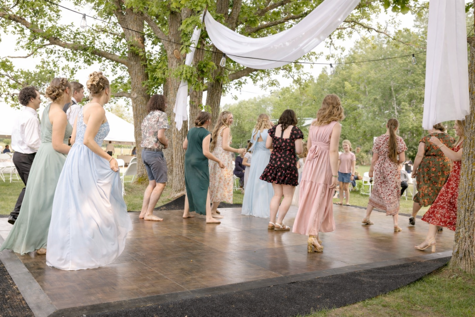 Group of women in dresses dancing on a wooden dance floor outdoors, decorated with white fabric and string lights, under tall trees, during a festive outdoor event.