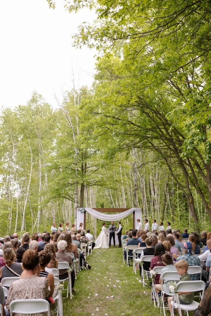 Outdoor wedding ceremony with a bride and groom exchanging vows under a wooden arch draped with white fabric, surrounded by guests seated on white chairs in a lush green forest setting.