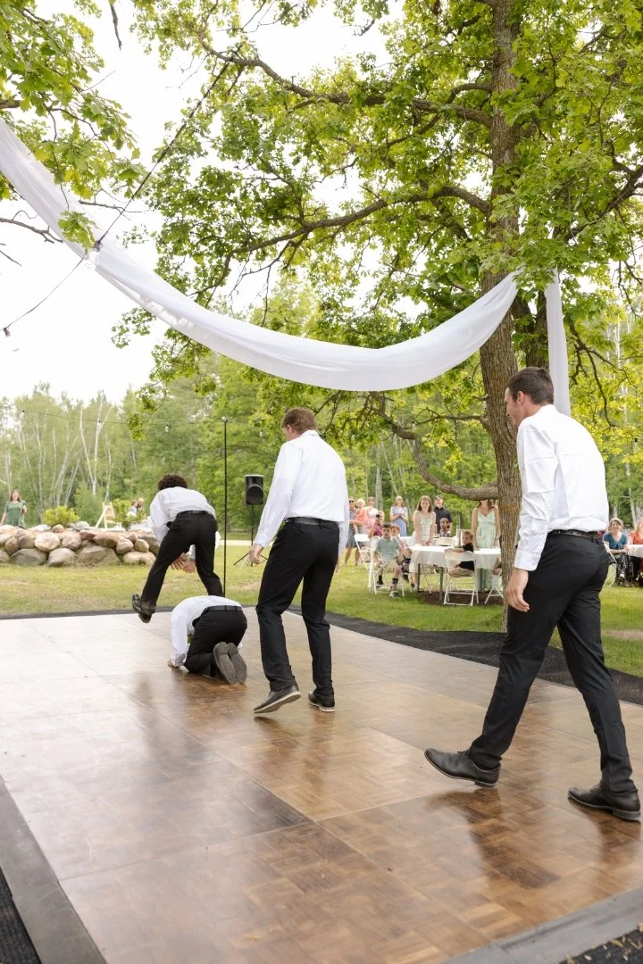 Groomsmen in white shirts and black pants at an outdoor wedding, with some standing and one kneeling, on a wooden dance floor under a large tree with green leaves, with guests seated at tables in the background.