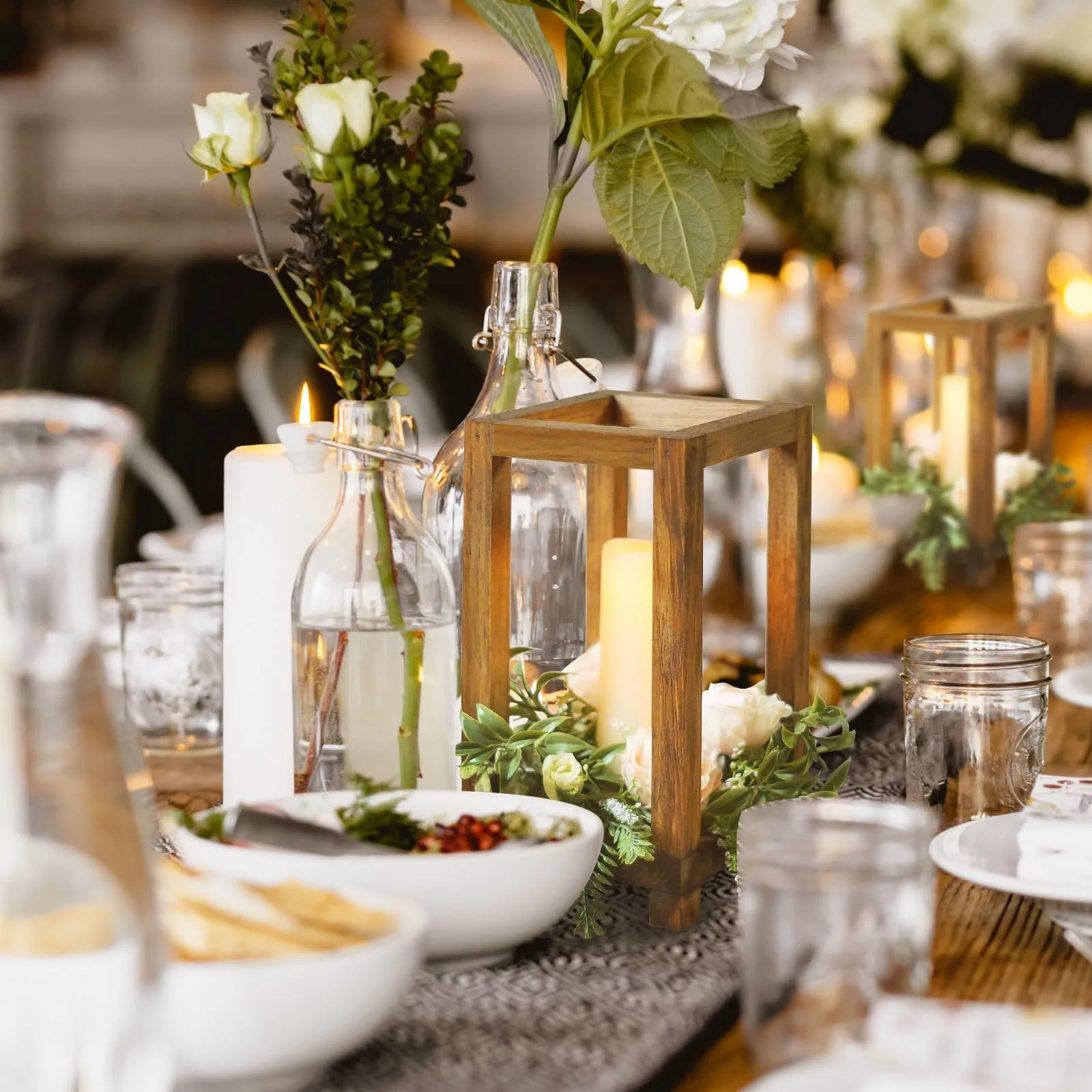 Table centerpiece with glass bottles holding white roses and greenery, wooden lanterns, candles, and a bowl of salad at an elegant event.