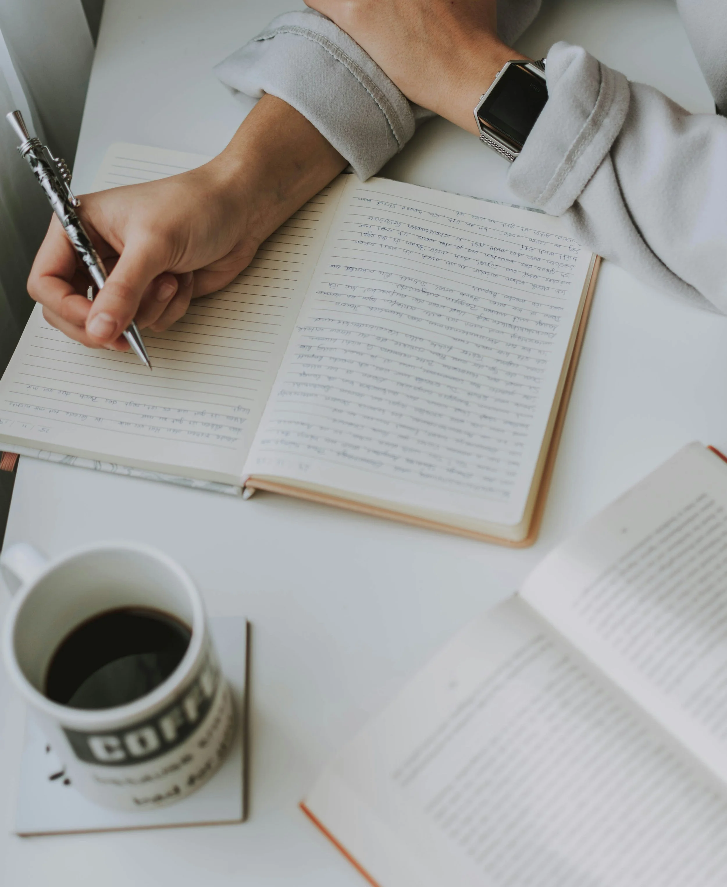 A person writing in a notebook with a pen, wearing a white sweater, with a smartwatch on their wrist, on a white table. There is an open book and a coffee mug on the table.