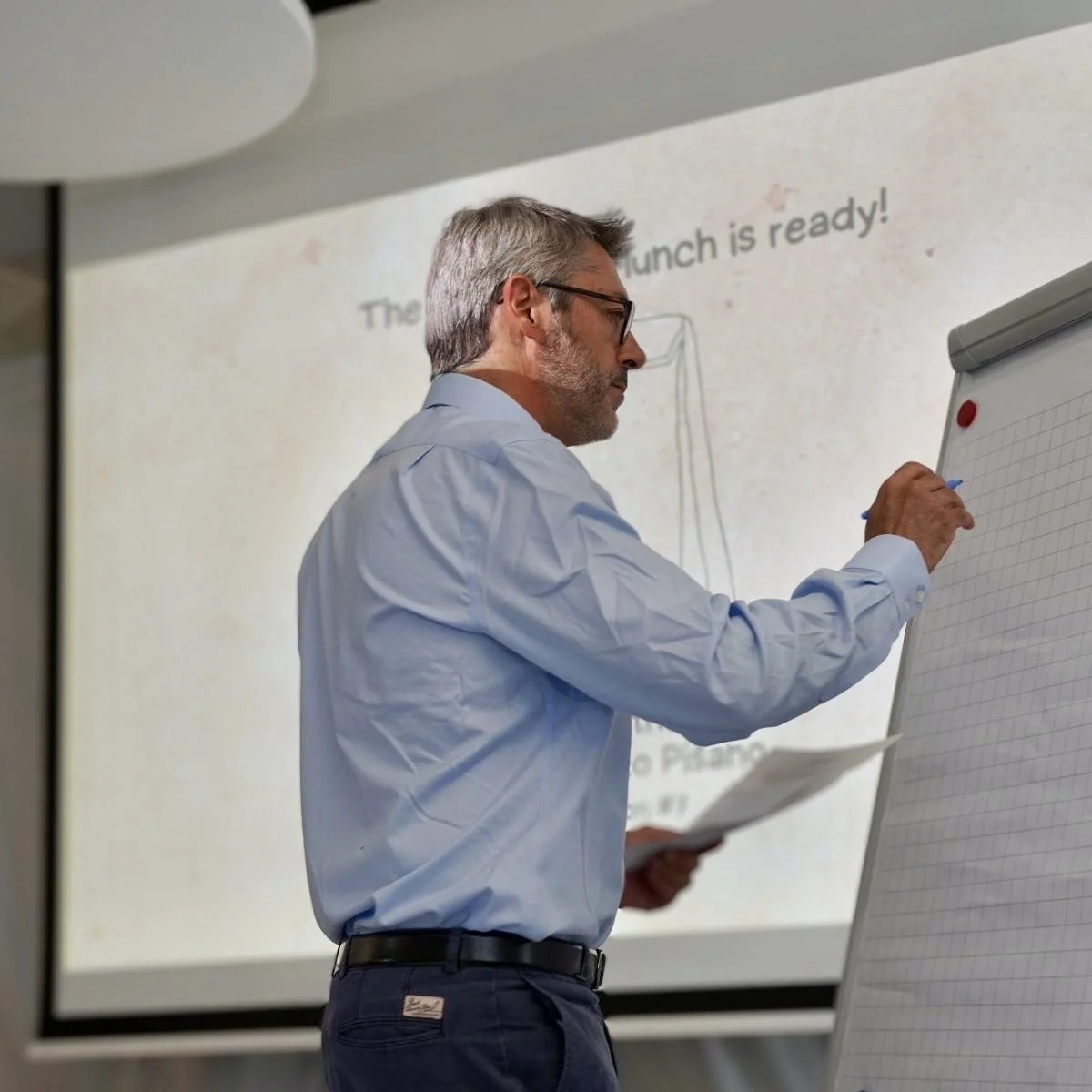 A man with gray hair and glasses writing on a flip chart during a presentation with a projected slide that says 'The lunch is ready!' in the background.