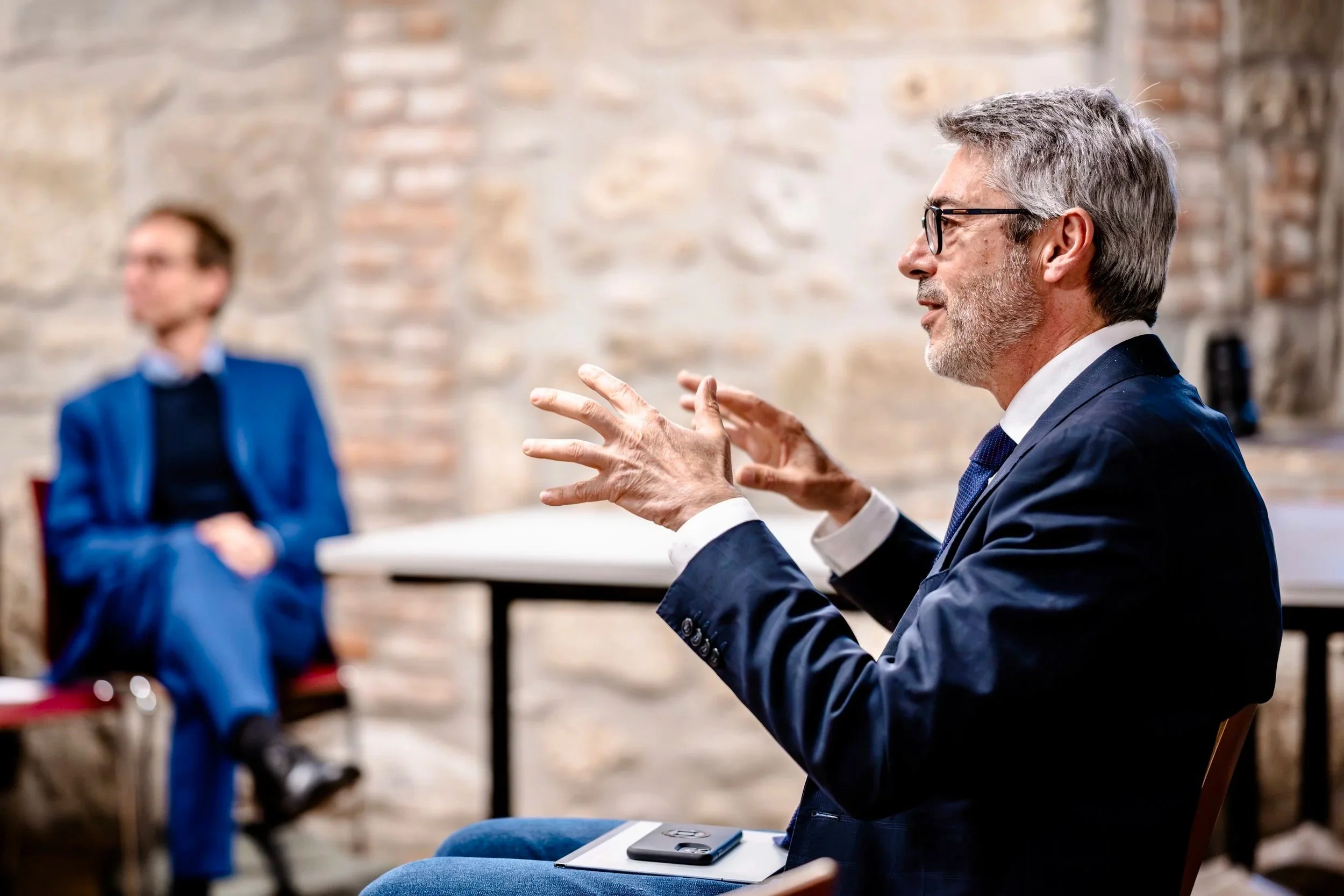 A man with gray hair, glasses, a white shirt, and a dark blue suit speaking and gesturing with his hands, giving a conference. A man in a blue suit sitting in the background in front of a brick wall.