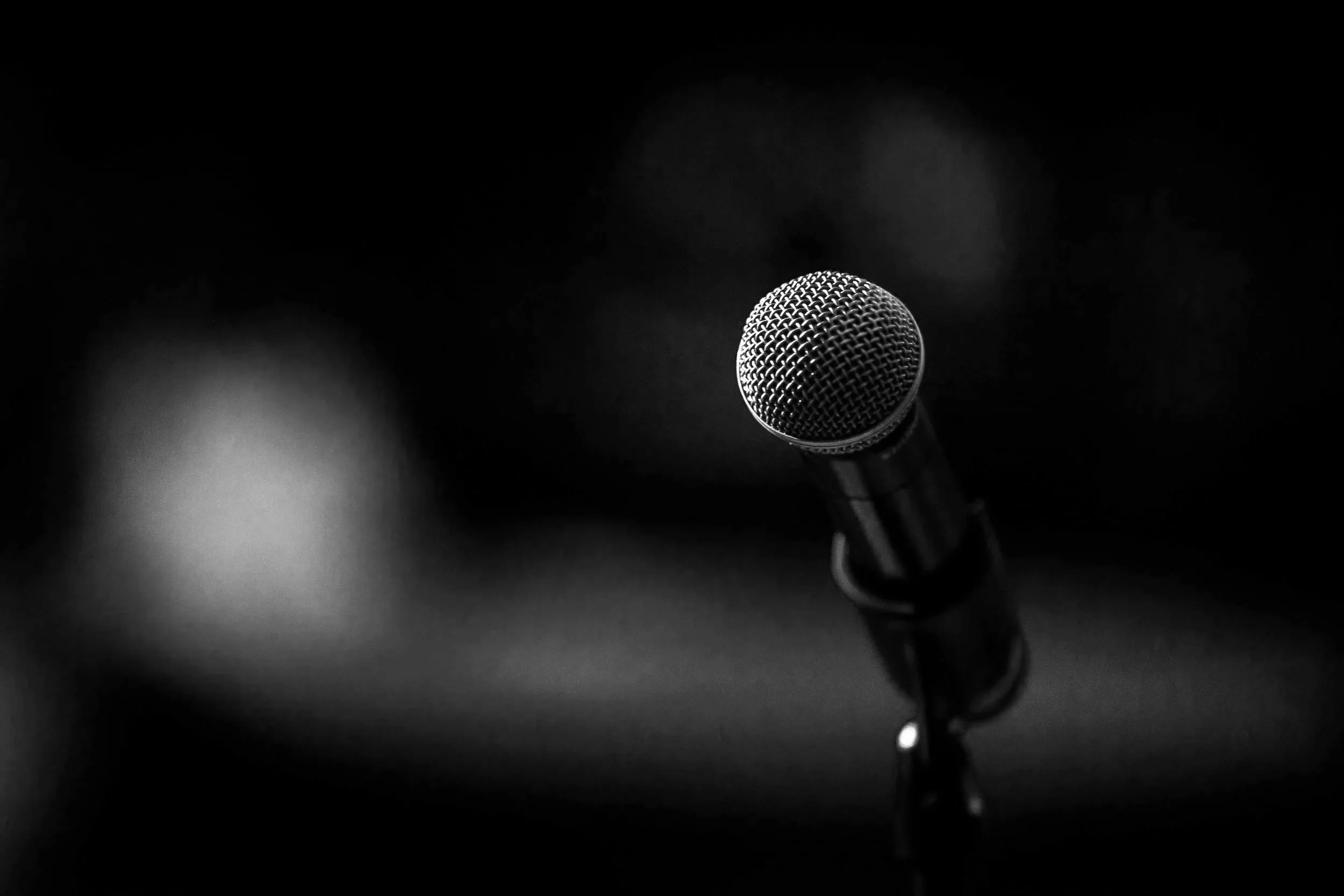 Black and white photo of a microphone on a stand in a dark room or stage.
