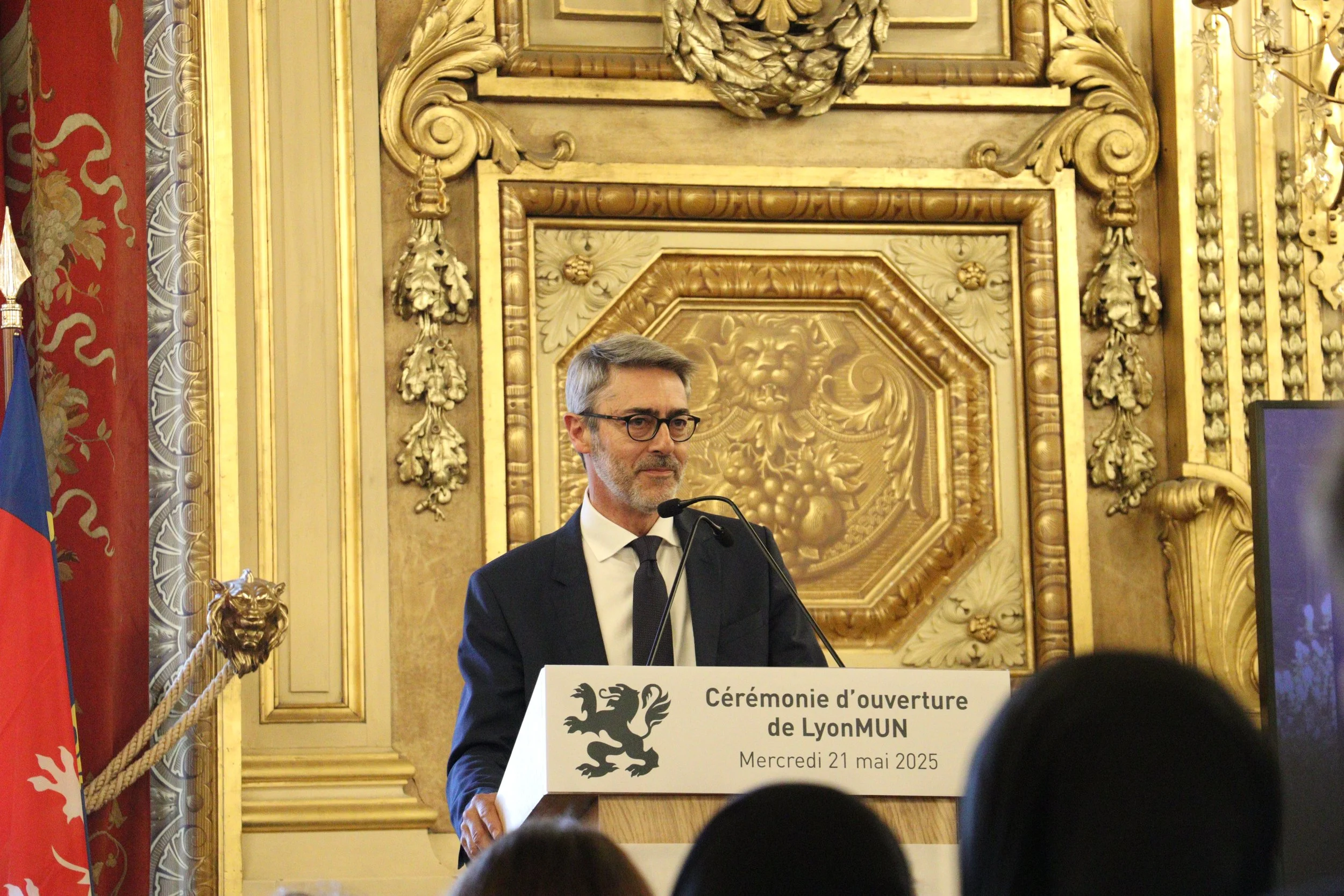 A man in a dark suit and tie, wearing glasses and sporting a beard, is standing at a podium delivering a speech during an event. The podium has a sign that reads "Cérémonie d'ouverture de LyonMUN Mercredi 21 mai 2025". The background features ornate gold detailing and decorations, with flags on the left side of the scene.