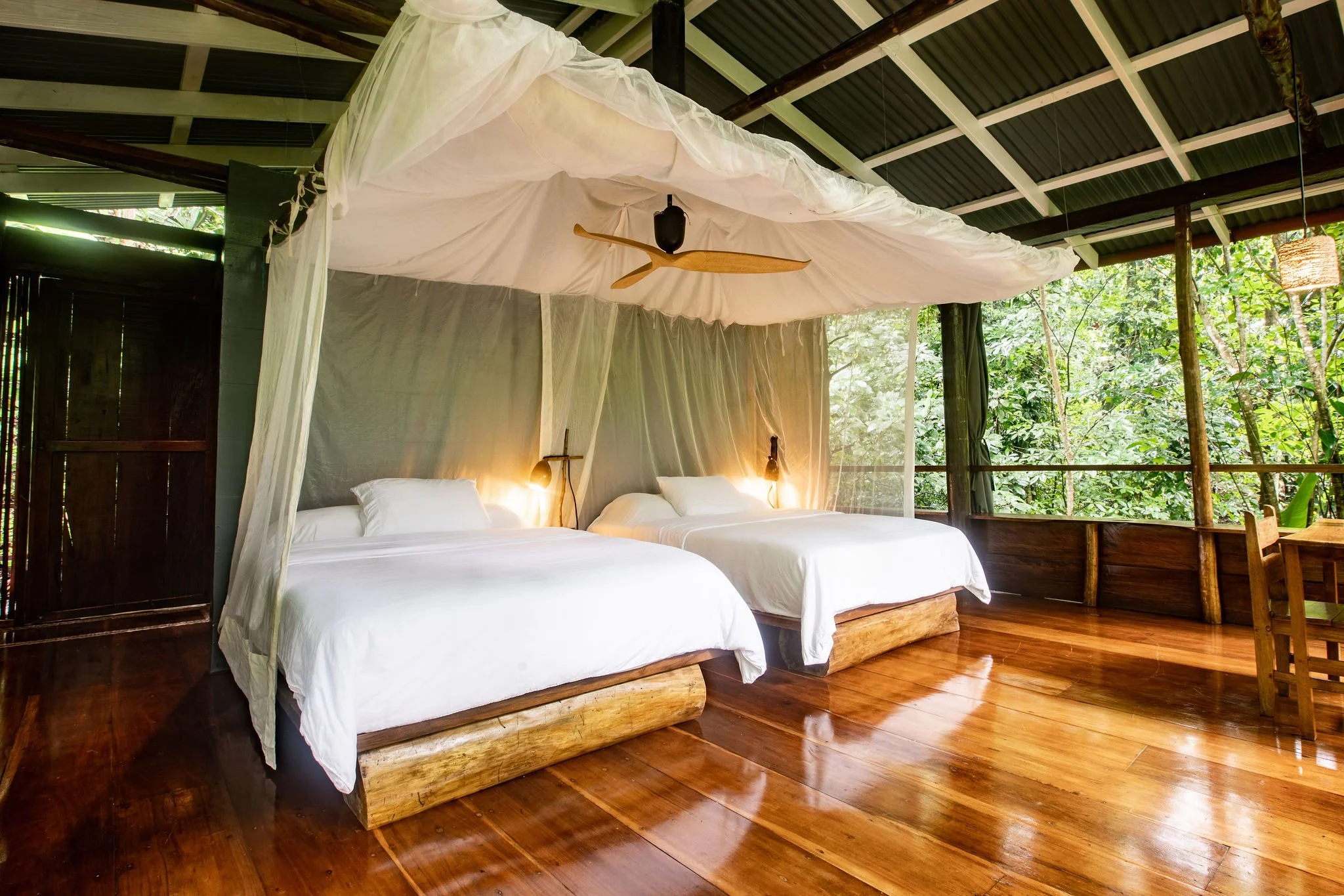 Two beds with white bedding in a rustic open-air room with wooden floors, canopy, and lush green trees outside.