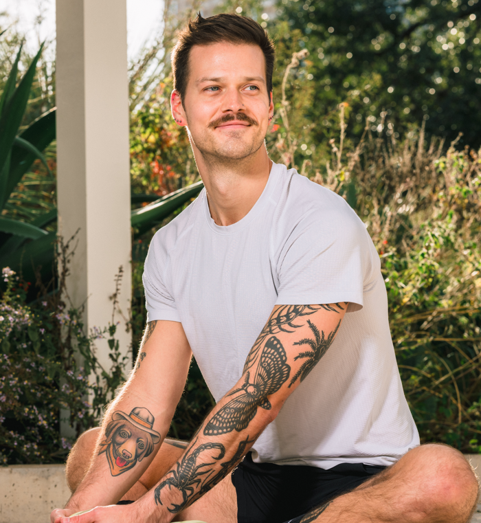 A young man with a mustache and tattoos sitting outdoors in a garden, wearing a white t-shirt and black shorts, looking to the side.