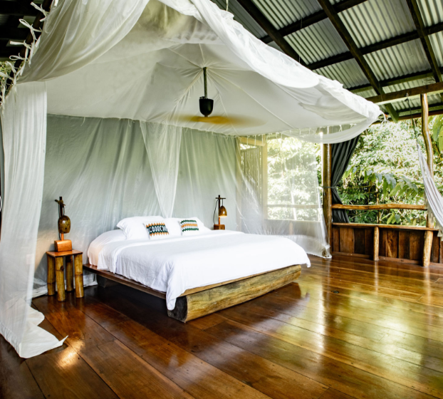 A rustic bedroom with a canopy bed, white bedding, wooden side tables, and natural surroundings visible through the open area.