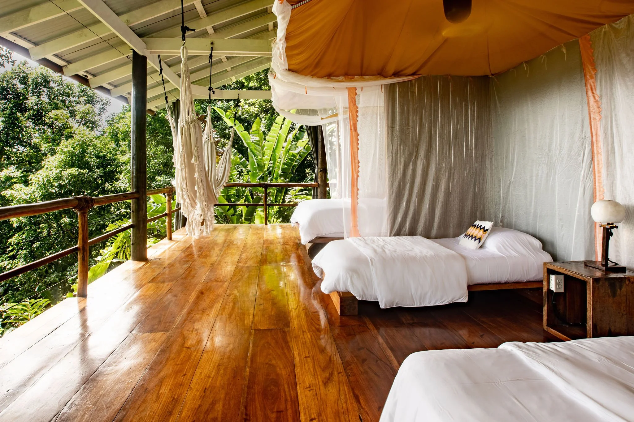 A cozy bedroom with white bedding and a decorative pillow, featuring a wooden side table with a lamp, and a canopy bed with sheer curtains, all opening to a balcony with lush green trees.