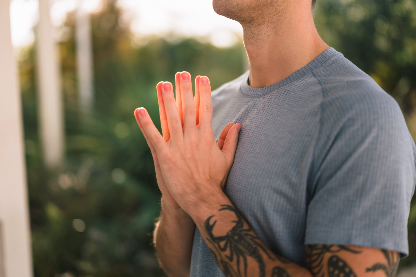 A person with a tattoo on their arm wearing a gray t-shirt, pressing their hands together on their chest in a prayer or meditative pose outdoors with blurred greenery in the background.