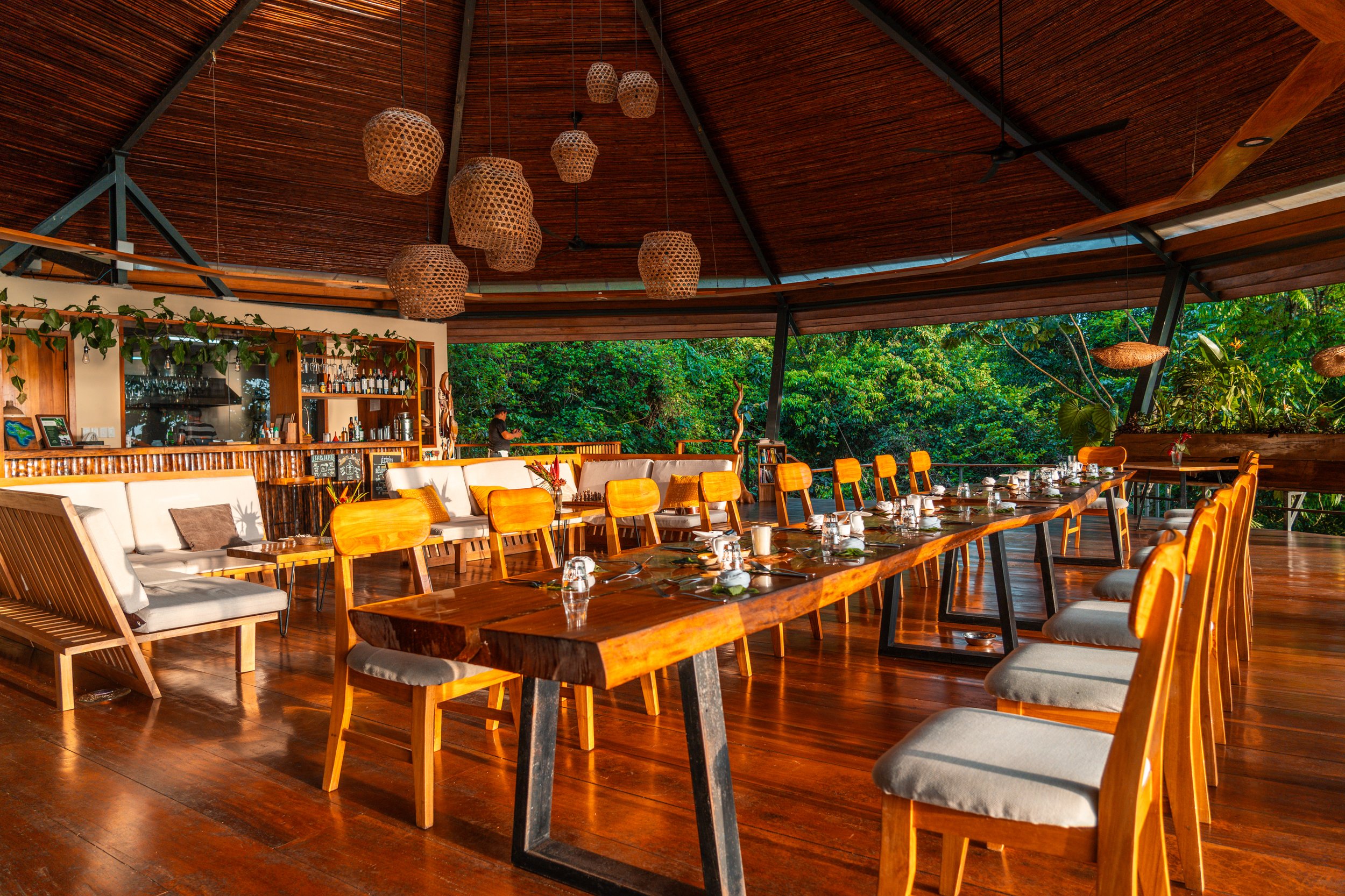 A spacious, open-air restaurant with a long wooden dining table and matching chairs, decorated with table settings and glasses, surrounded by lush greenery and hanging woven lanterns from a wooden ceiling.