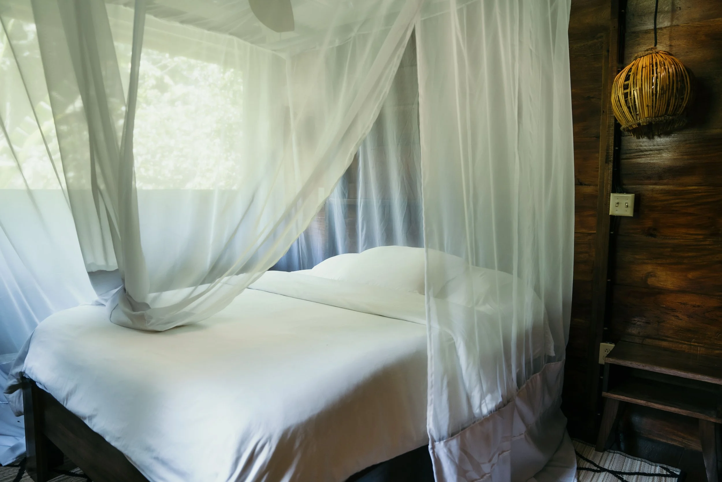 A bedroom with a bed enclosed by sheer white curtains, a wooden side table, a woven hanging lamp, and a window with white curtains and green foliage outside.