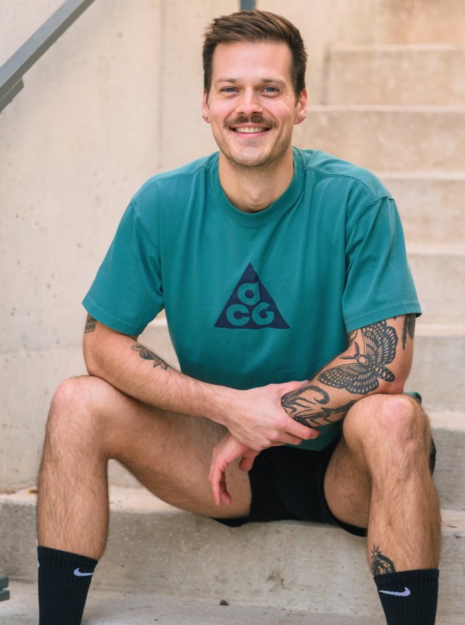 A young man with short brown hair, a mustache, and tattoos on his arms, sitting on concrete stairs with a smile. He's wearing a teal T-shirt with a triangle logo, black shorts, and black Nike socks.