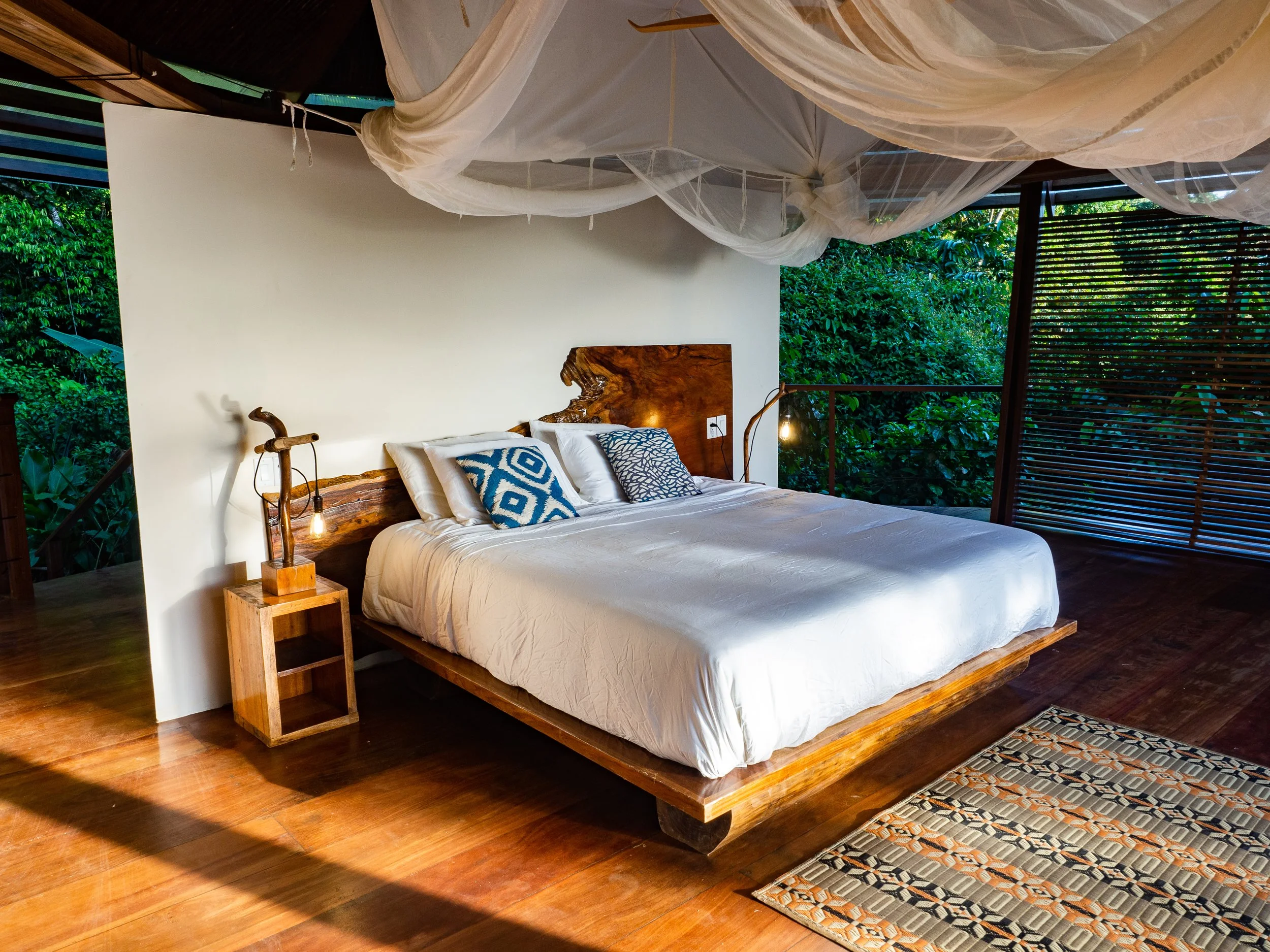 A rustic bedroom with a wooden bed frame, white bedding, patterned pillows, two bedside tables with small lamps, a window with greenery outside, and a wooden floor with a patterned rug. A sheer canopy drapes from the ceiling.