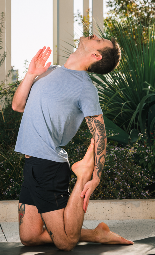 A man practicing yoga outdoors, performing a backbend with one leg lifted and foot held by his hand, near greenery and a white fence.