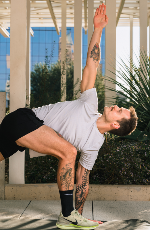 Young man performing a yoga pose outdoors, with tattoos on his arm and leg, under a structure with a modern building in the background.