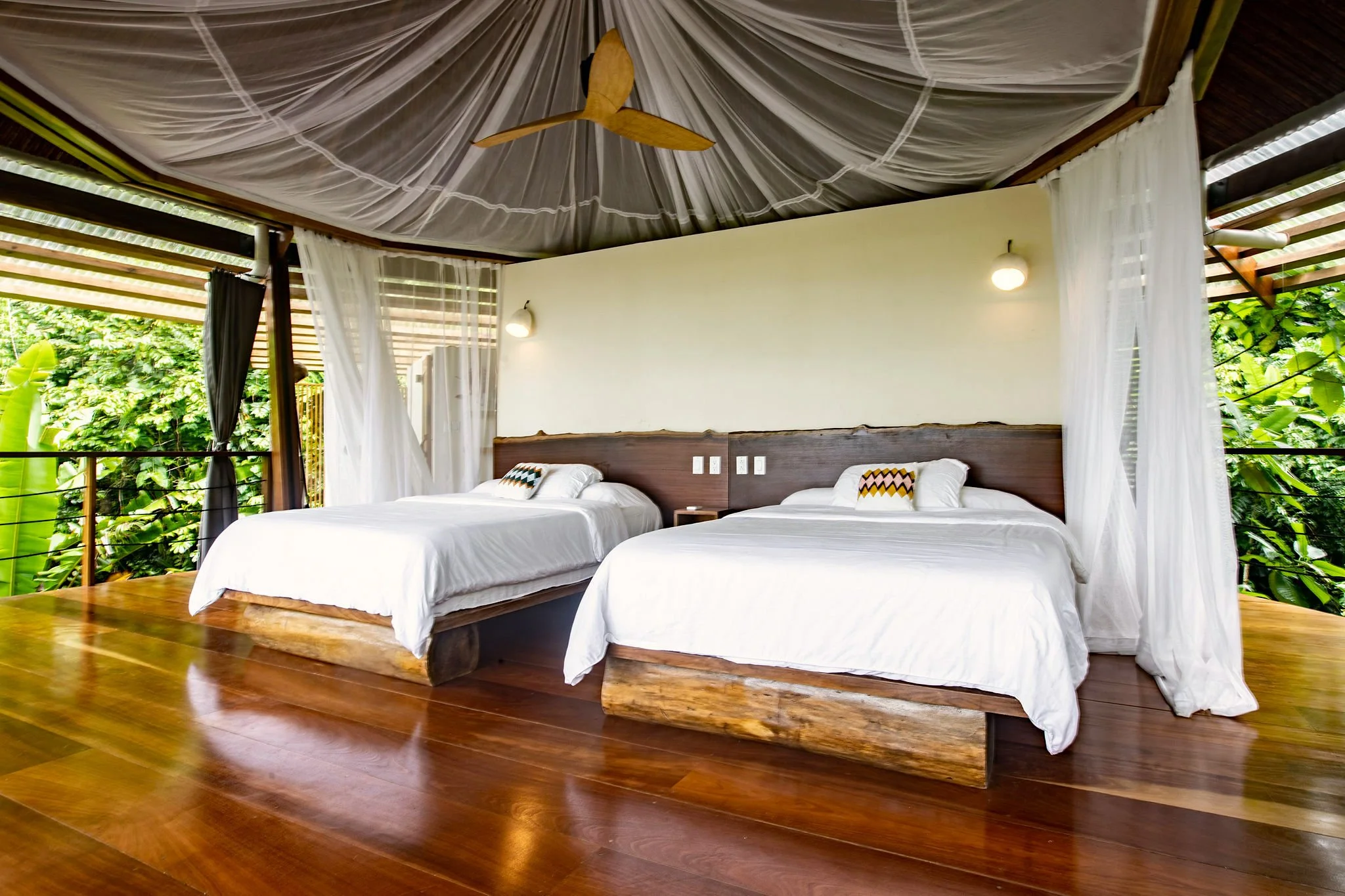 Two double beds with white linens in an outdoor bedroom with wooden floors, surrounded by lush green foliage, white curtains, and a ceiling fan.