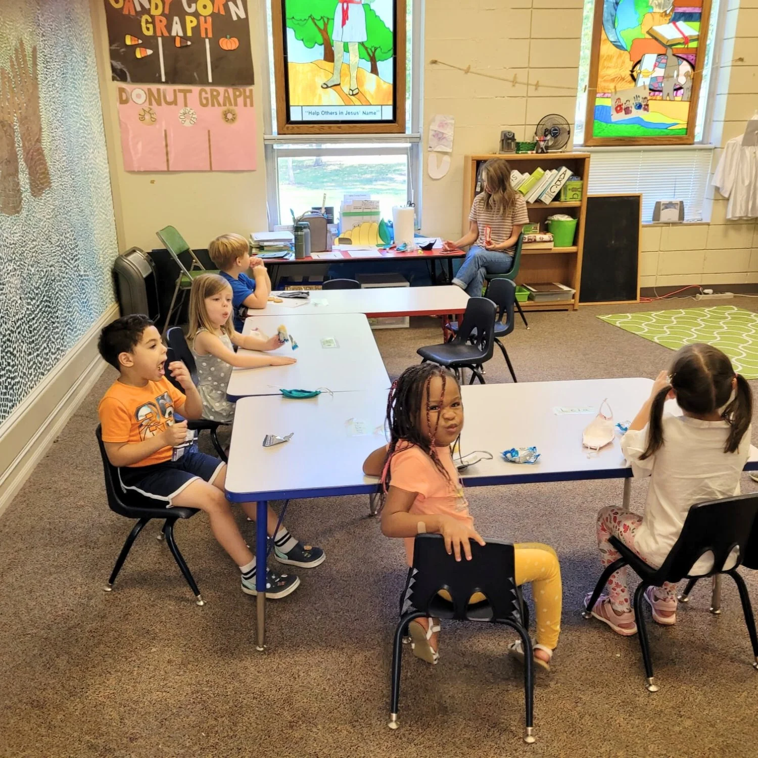 A classroom with six young children sitting at tables, some with masks on the table in front of them. There is a teacher or caregiver at the back of the room, sitting at a desk. The classroom has colorful children's artwork on the walls and windows, with educational posters and bookshelves.