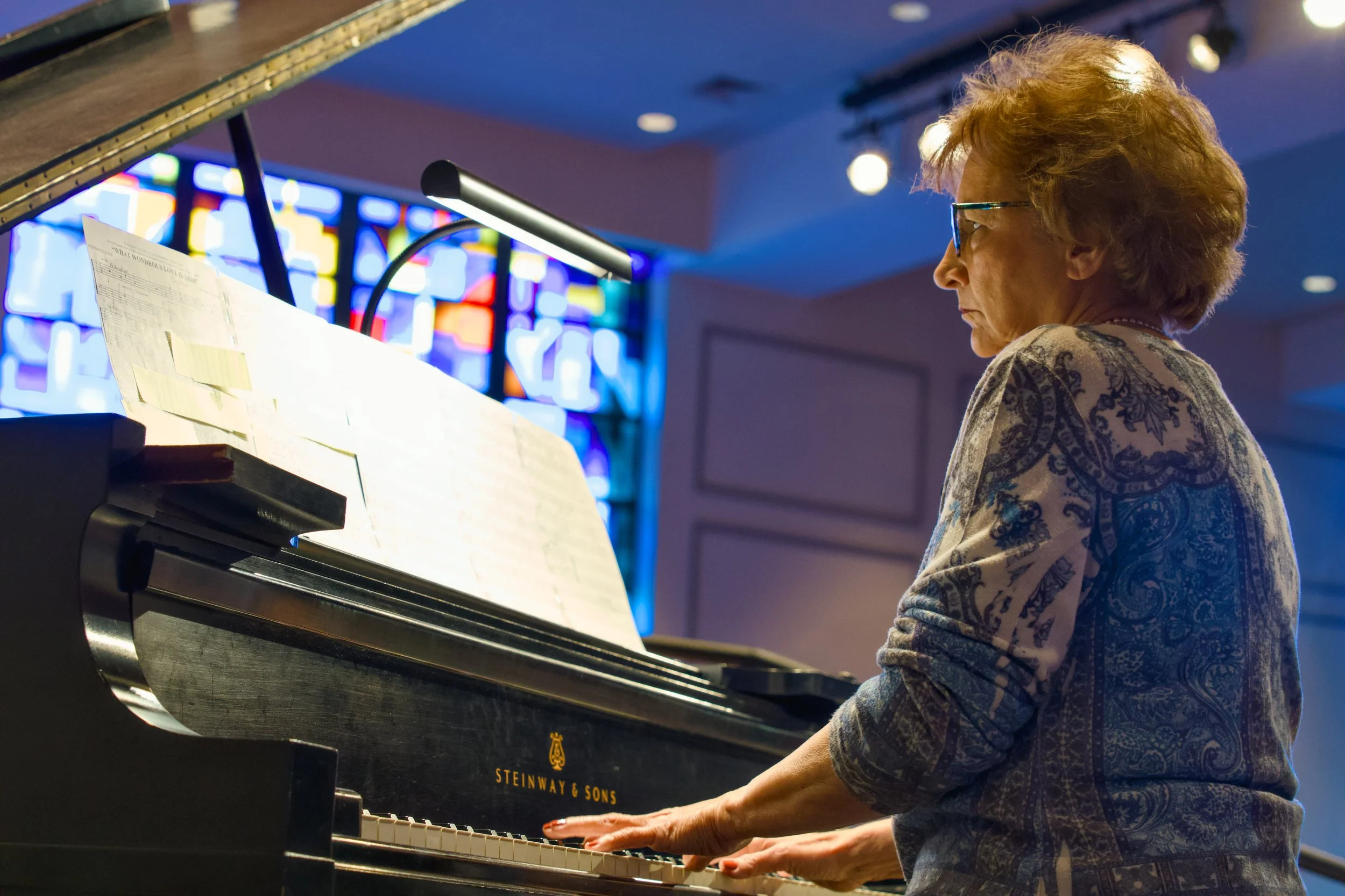 A woman with short red hair and glasses playing a black Steinway & Sons grand piano, with sheet music on the music stand, in a room with a stained glass window in the background.