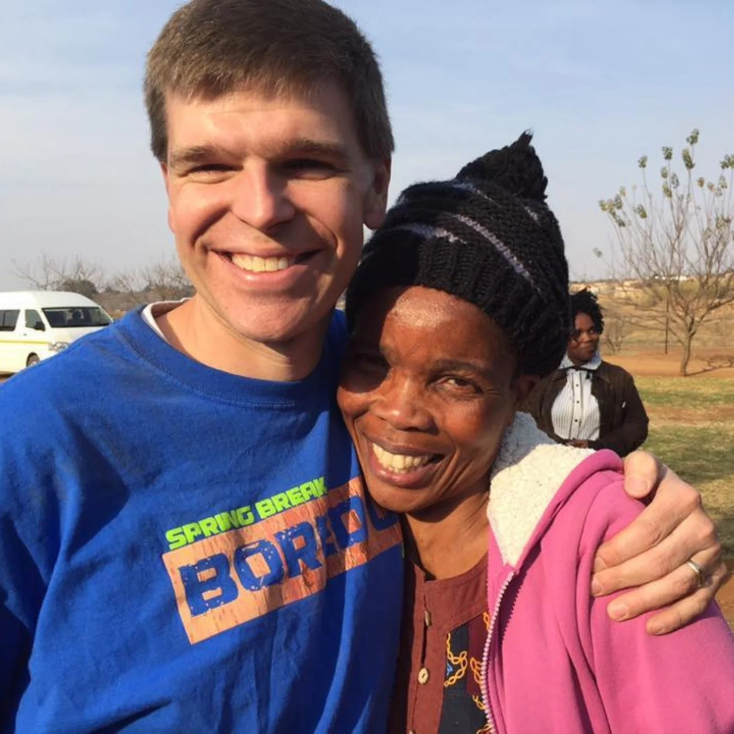 A young man in a blue shirt smiling and hugging an older woman in a pink jacket and black beanie, both outdoors on a sunny day.