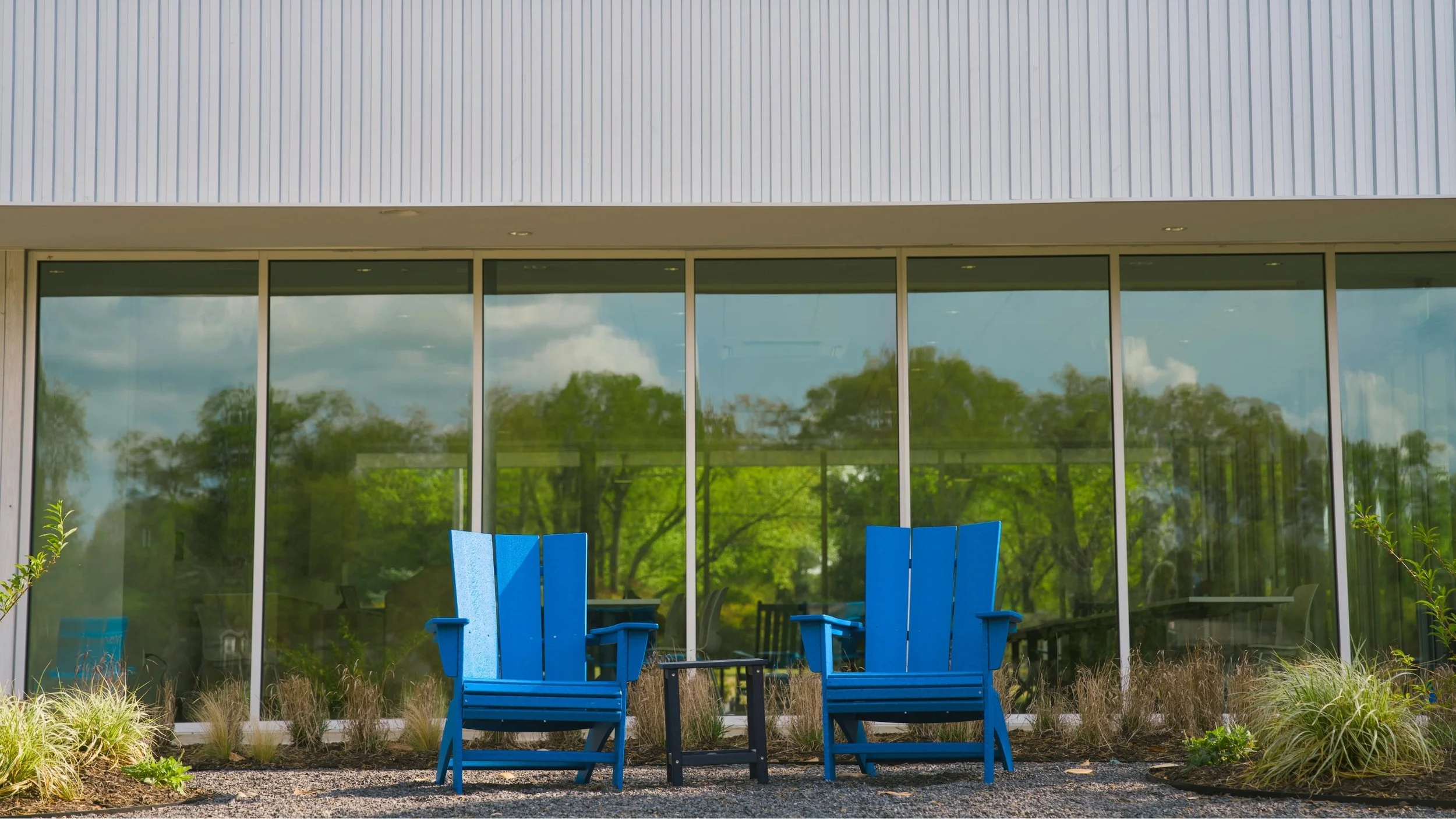 Two blue Adirondack chairs with an empty black table between them in front of a large glass window reflecting green trees and a cloudy sky.