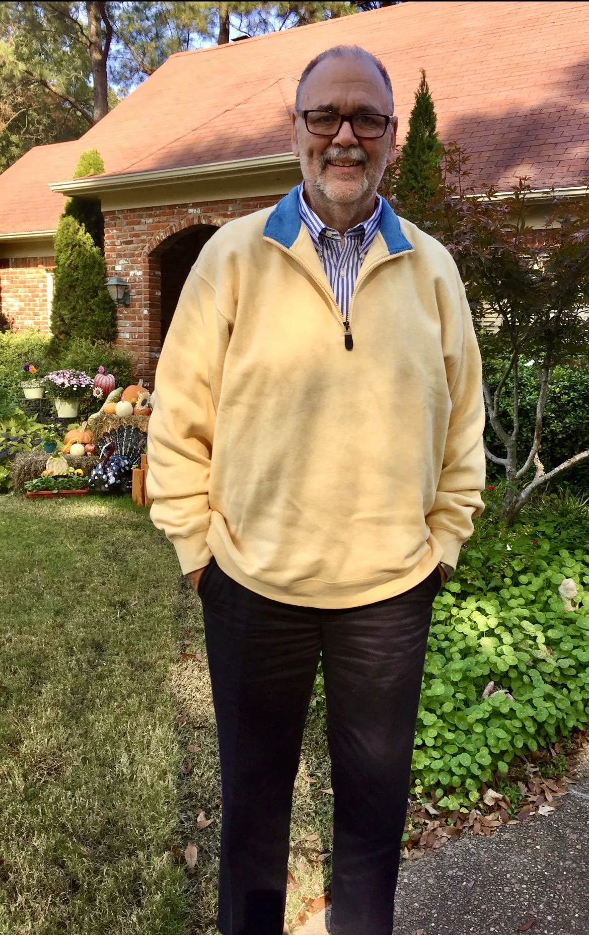 A smiling man with glasses and a beard standing outside in front of a brick house with a red shingle roof. He is wearing a yellow fleece pullover and dark trousers, with his hands in his pockets. There are flowers and decorative pumpkins on the lawn behind him.
