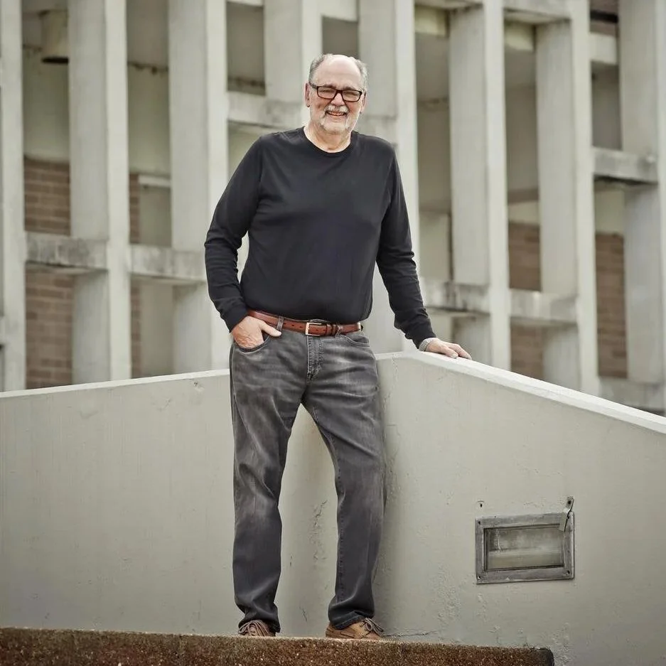 A smiling elderly man with glasses, wearing a black long sleeve shirt and gray jeans, standing outdoors on a concrete balcony wall, with a modern building in the background.