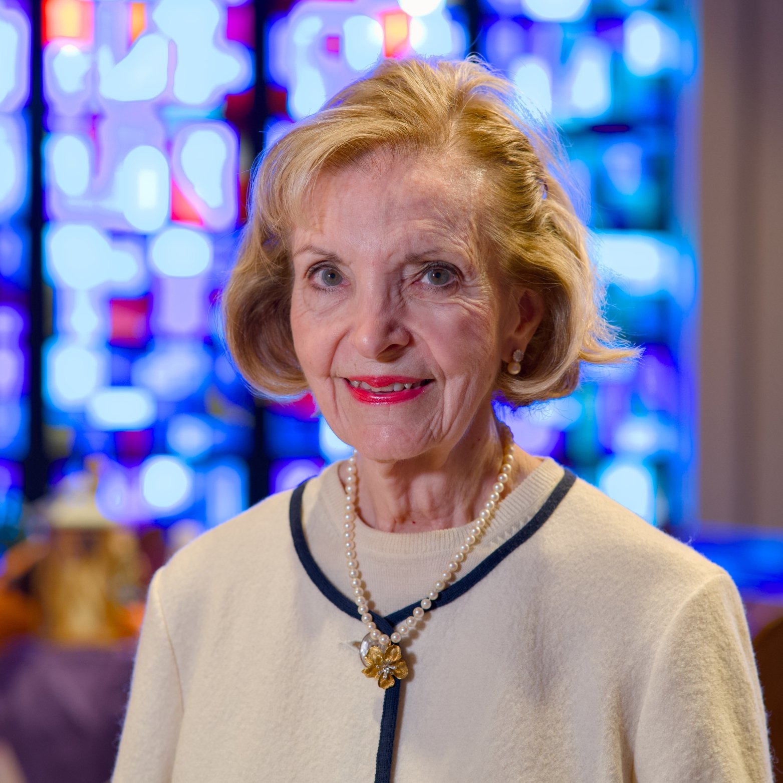 An elderly woman with short blonde hair, wearing a pearl necklace, earrings, and a cream-colored sweater with navy trim, standing in front of colorful stained glass windows.