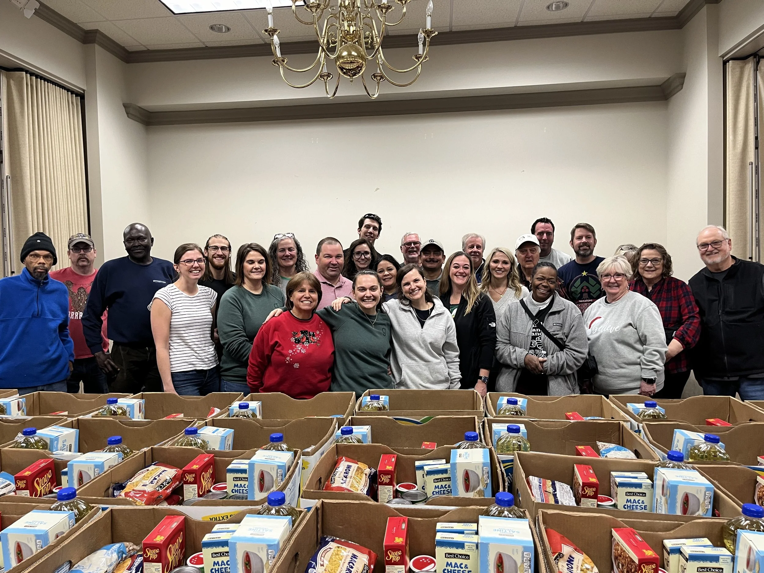 Group of people standing behind boxes of food donations in a room with a chandelier and beige walls.