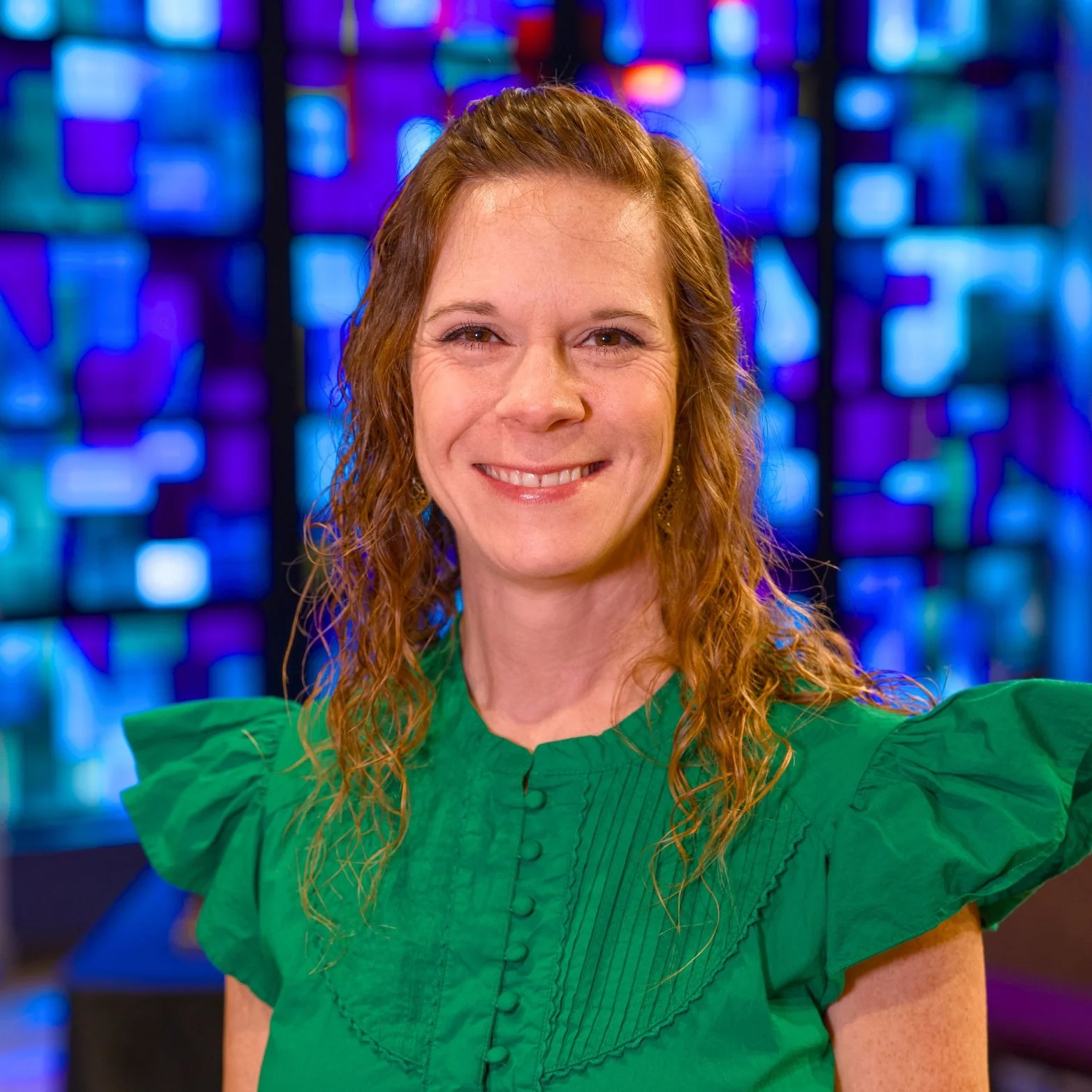 A woman with wavy red hair wearing a green top with ruffled shoulders, smiling, with a colorful, abstract, blue and purple background.