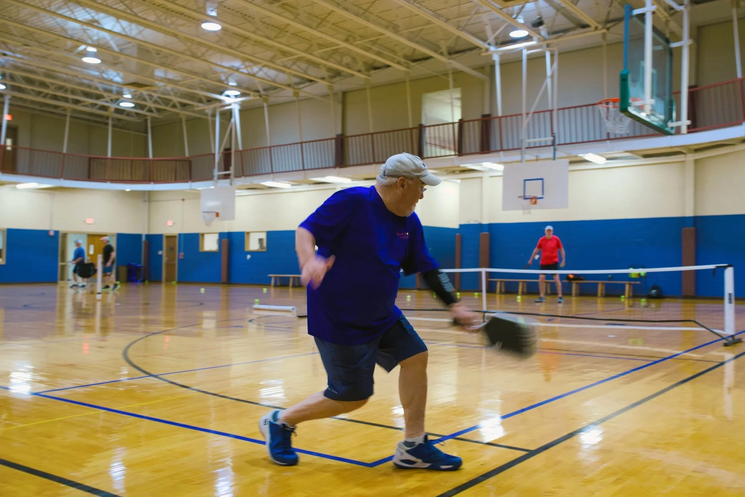 Older man playing paddle tennis inside a gymnasium with other players in the background.