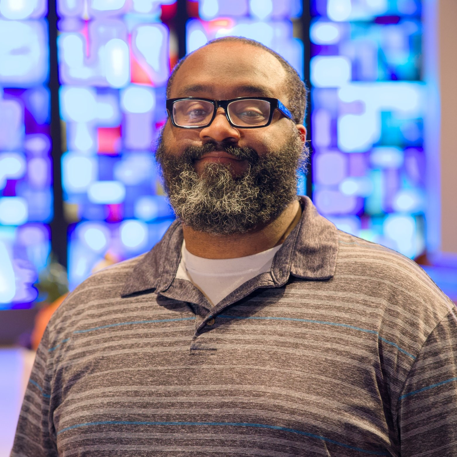 A man with glasses and a beard with gray streaks, wearing a striped polo shirt, standing indoors with a brightly colored stained glass window in the background.