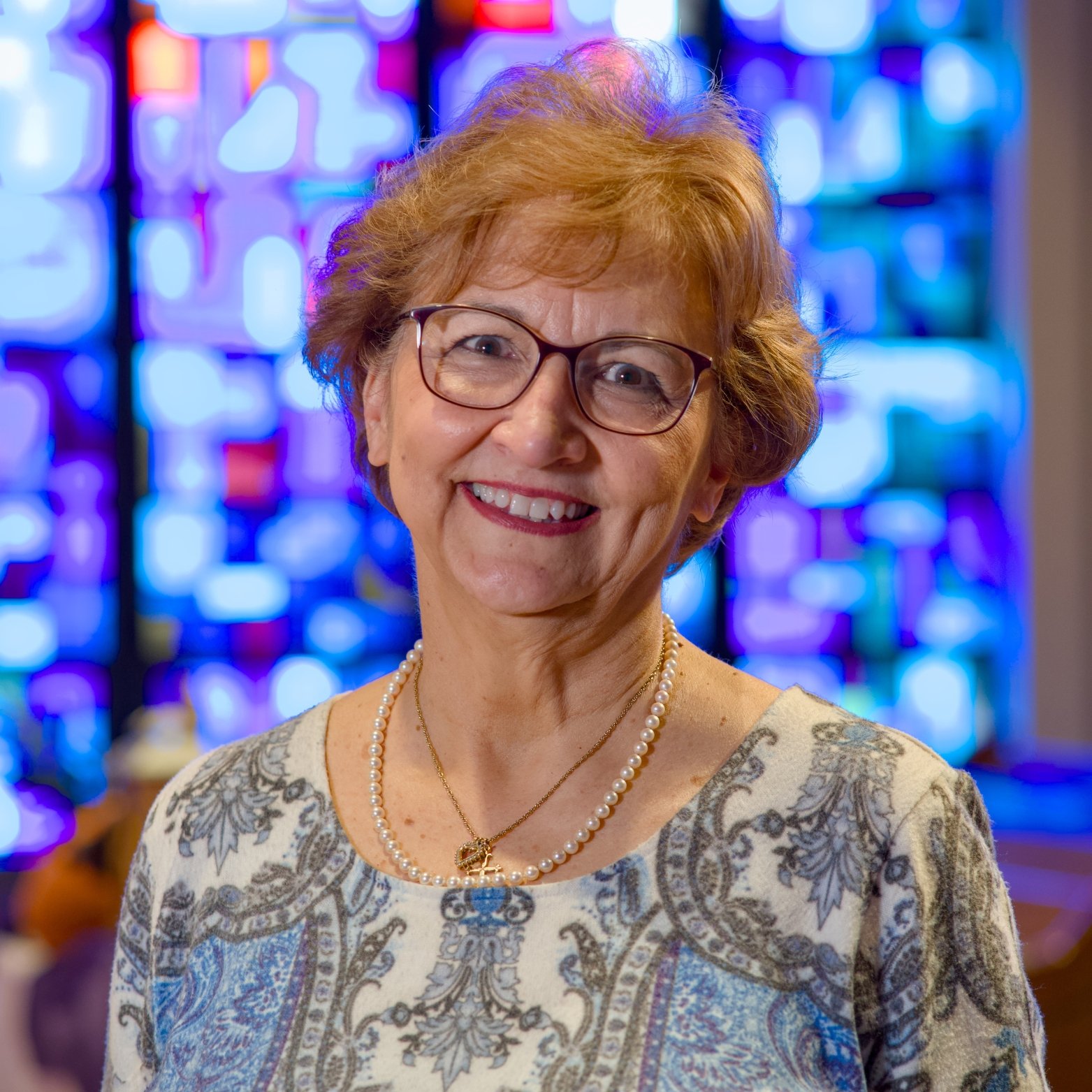 A smiling elderly woman with short curly hair, wearing glasses, a patterned blouse, pearls, and a gold necklace, standing in front of a colorful stained glass window.