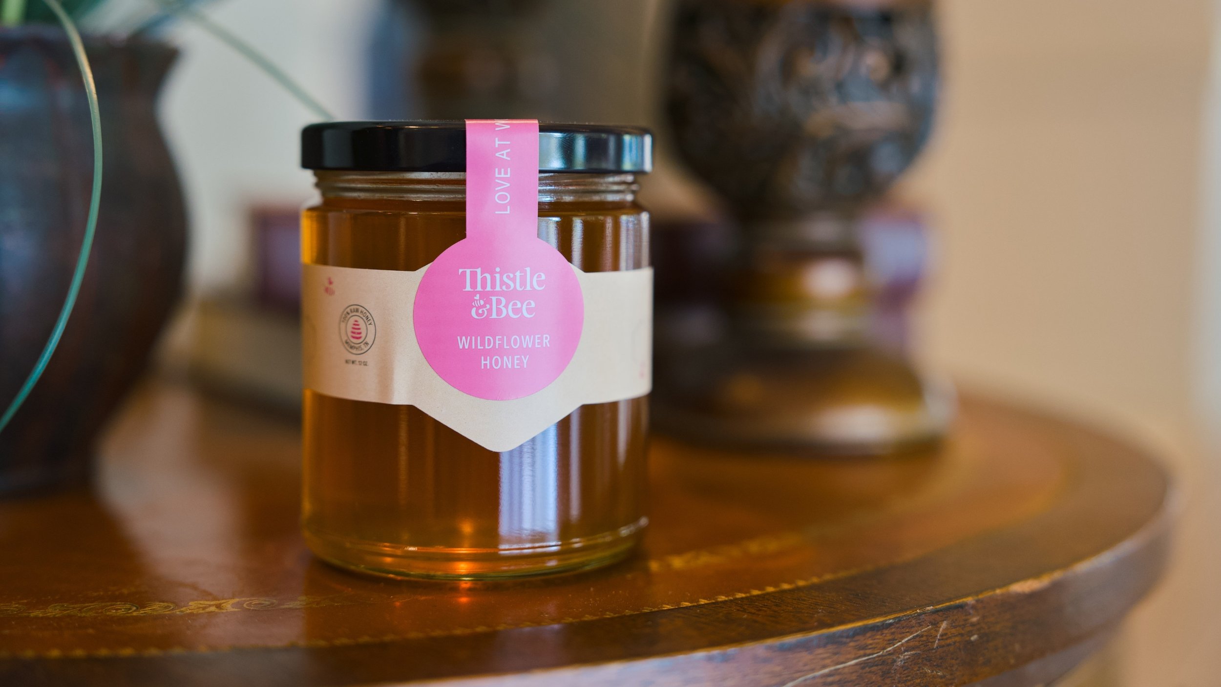 A jar of Thistle & Bee wildflower honey on a wooden surface, with decorative pottery in the background.