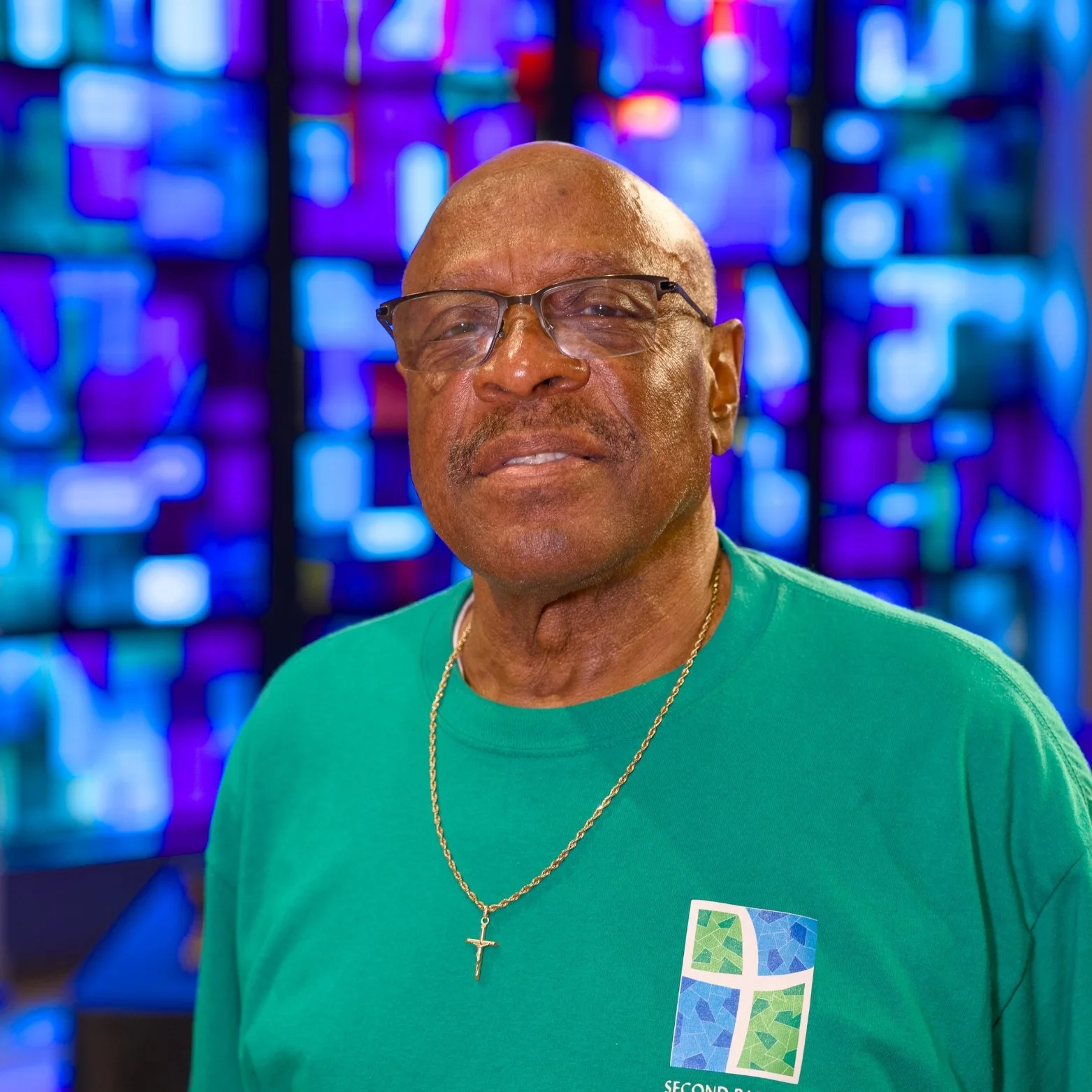 A man wearing glasses, a green t-shirt with a logo, a gold cross necklace, standing in front of a colorful stained glass window.