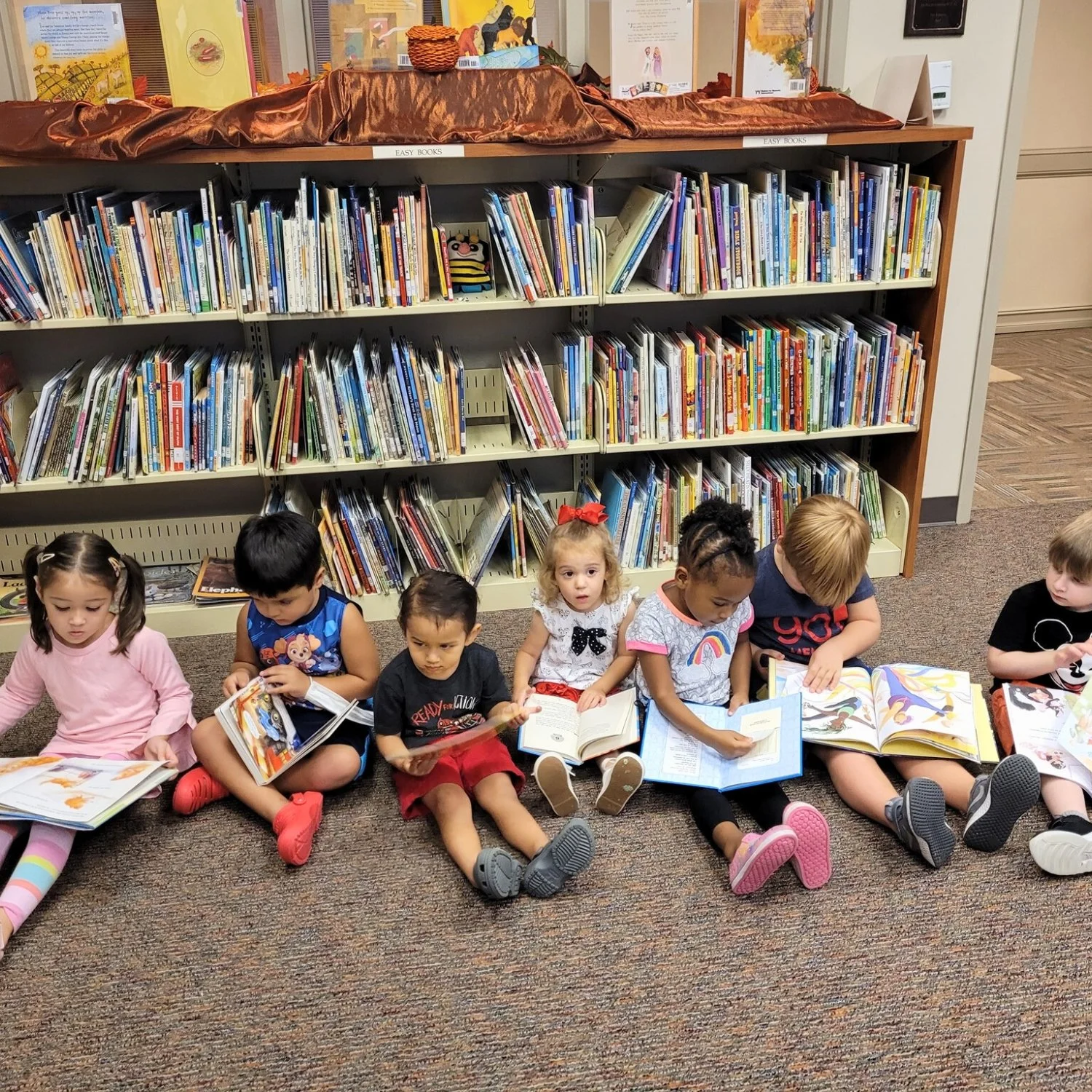 Seven young children sit on the floor in a library, reading books in front of a bookshelf filled with children's books.