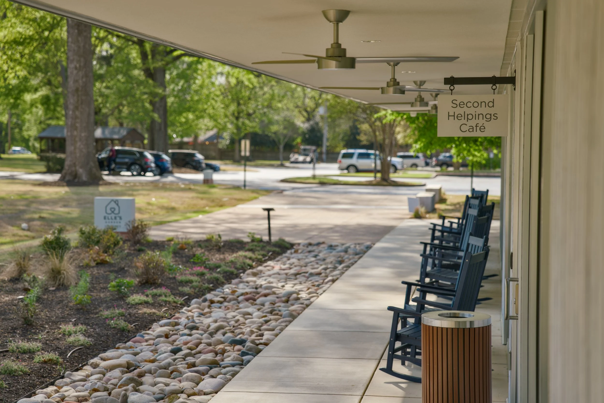 A sidewalk with a row of black rocking chairs under a beige awning, next to a garden with small plants and rocks, and a sign that reads 'Second Helpings Café.'