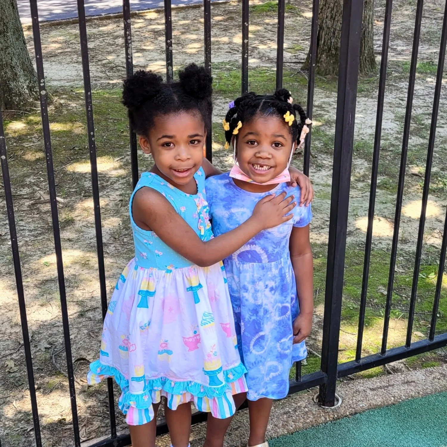 Two young girls in colorful dresses standing in front of a black metal fence, smiling, with one girl having her arm around the other.