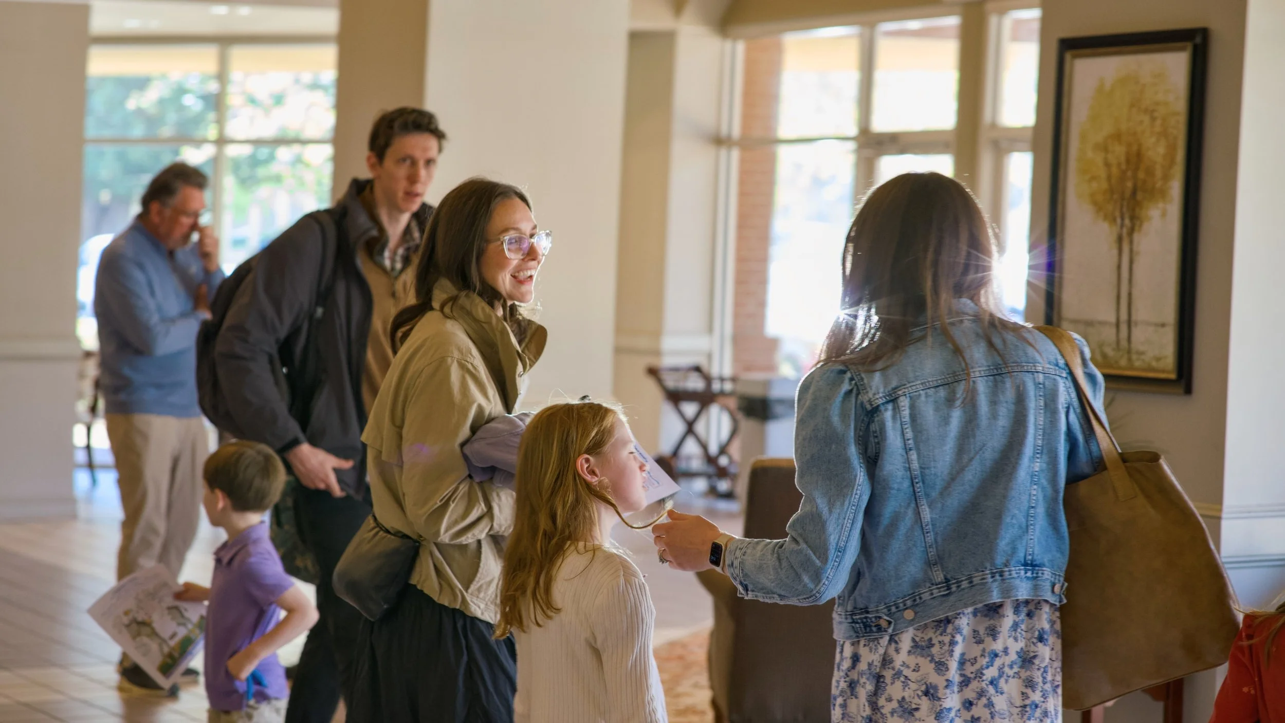 People standing in a lobby, engaging in conversation. A woman with glasses and a girl are in focus, talking to another woman in a denim jacket. Other people are in the background, some holding papers.