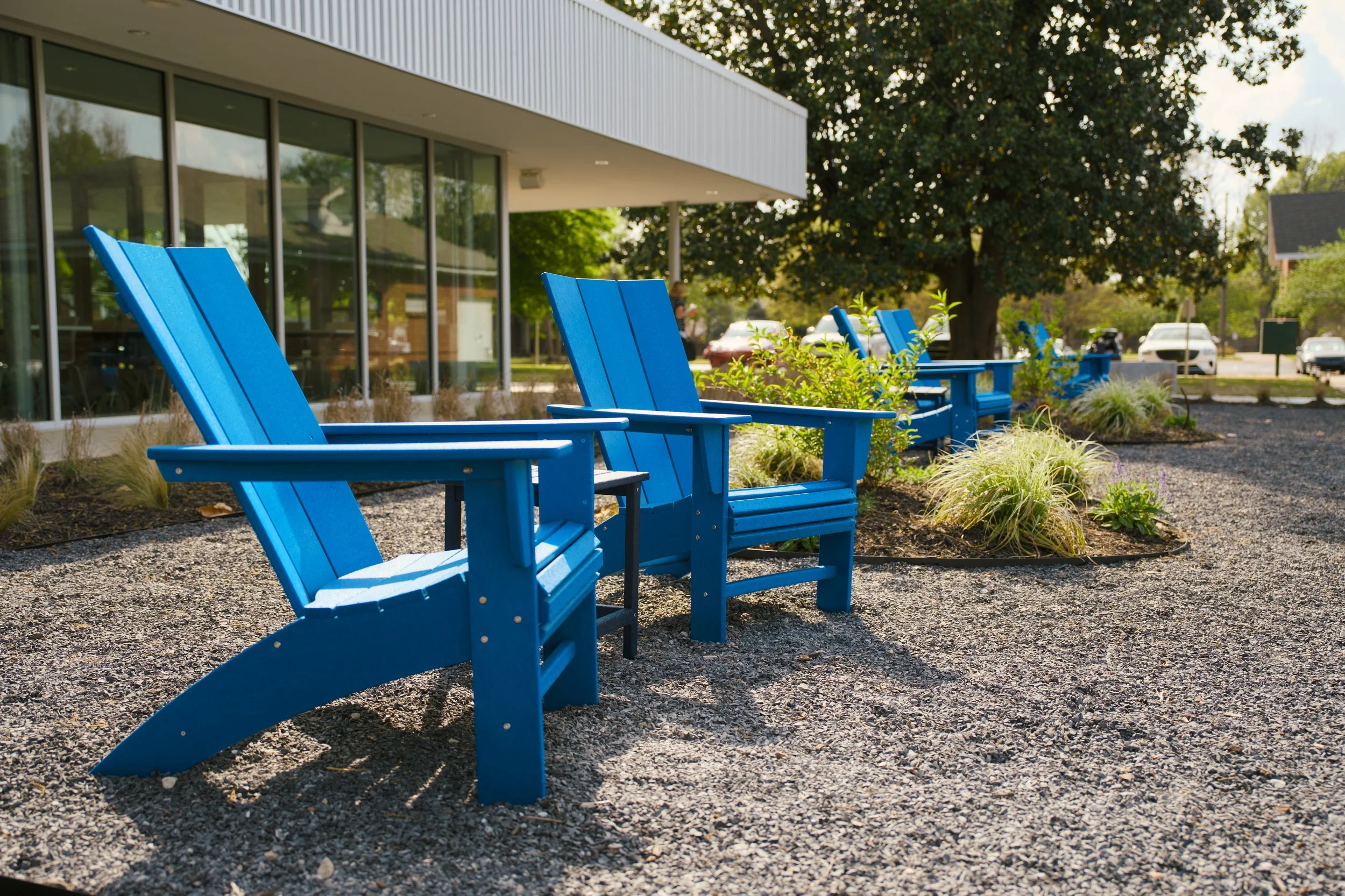 A row of bright blue wooden chairs on a gravel pathway outside a modern building with large glass windows, green plants, and trees in the background.